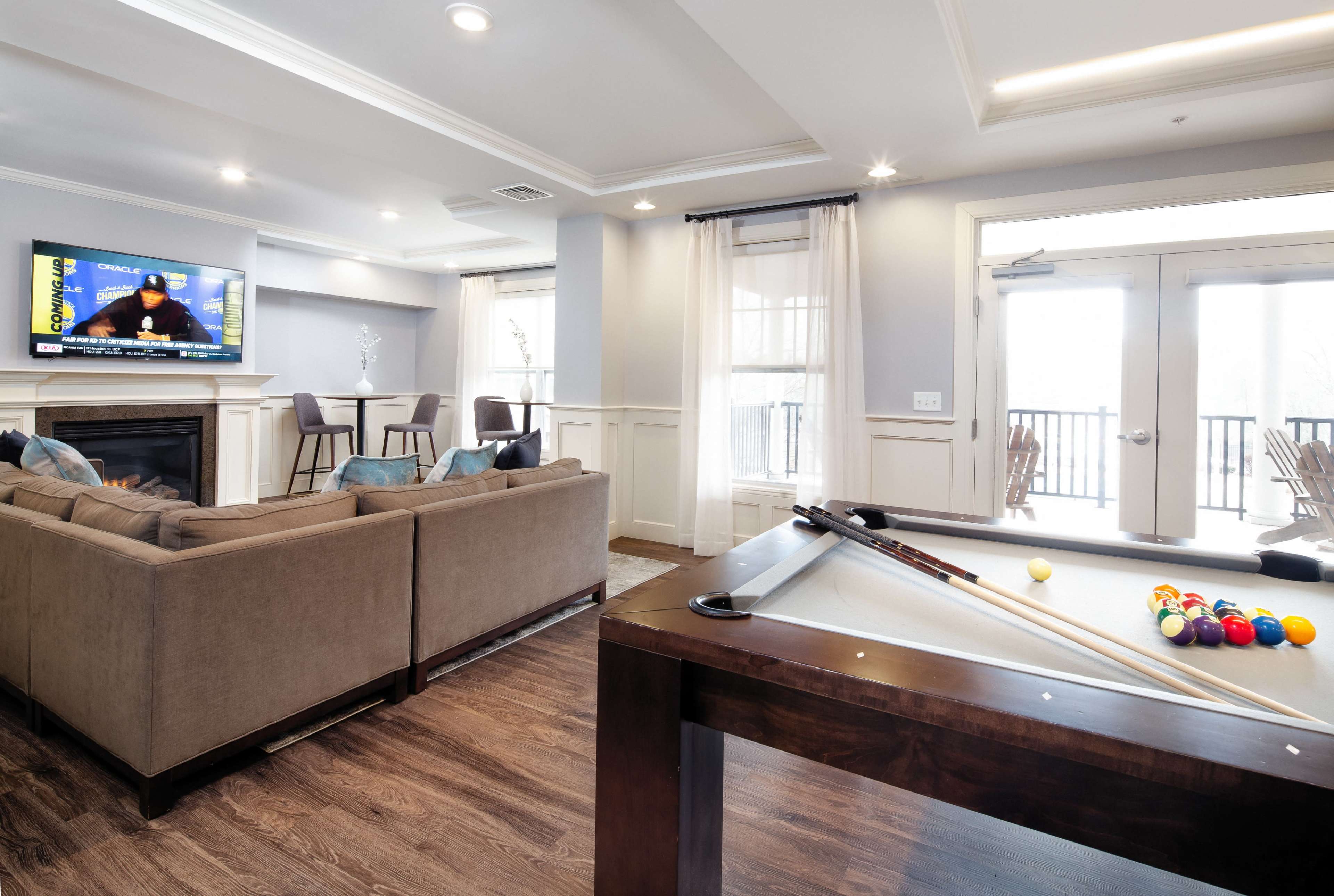 A modern living room with a pool table, a cozy sofa facing a television mounted above a fireplace, and large windows leading to a balcony.