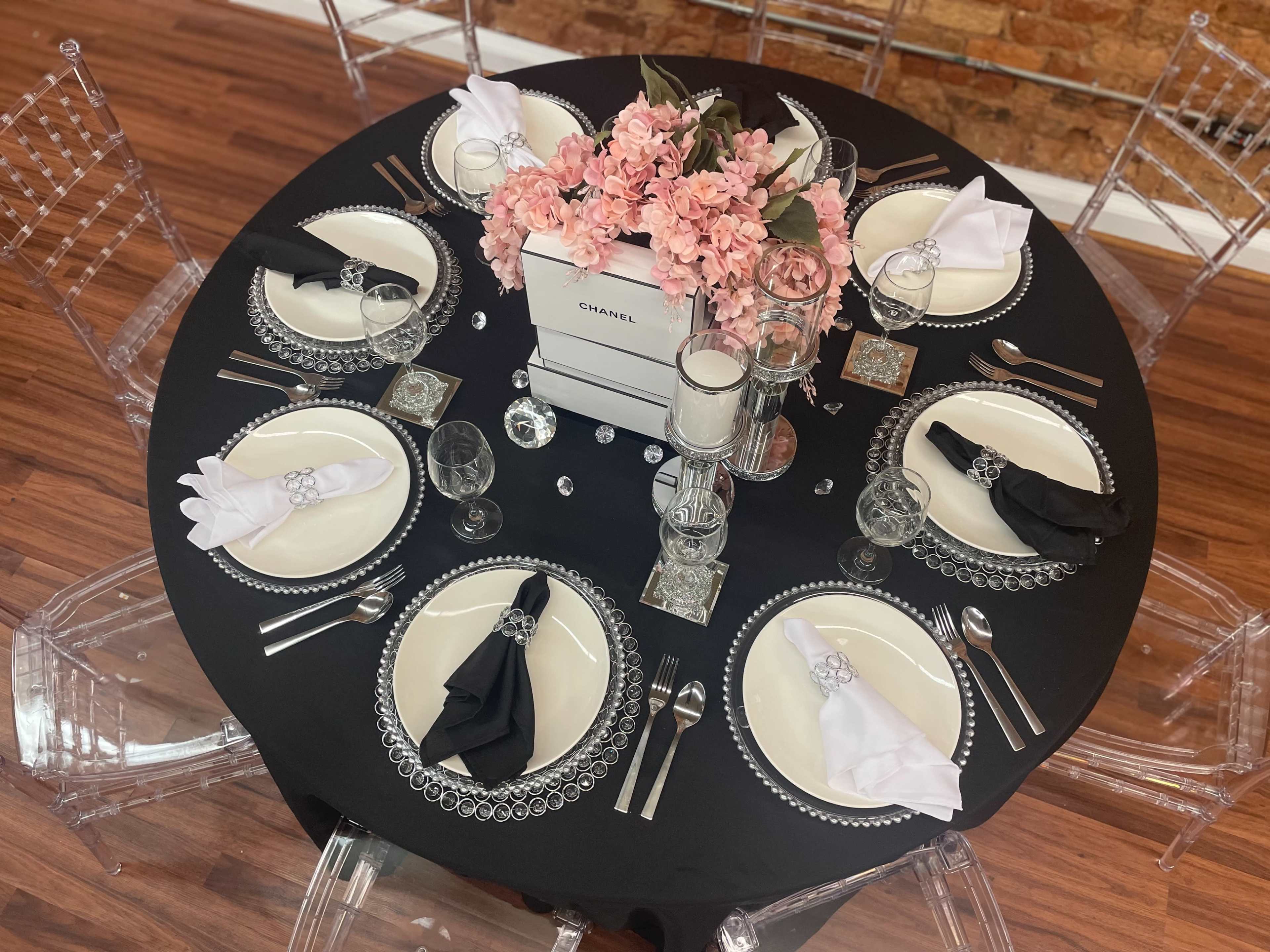 A round black table is elegantly set with white and black plates, silver cutlery, crystal glassware, and a pink floral centerpiece in a Chanel box.