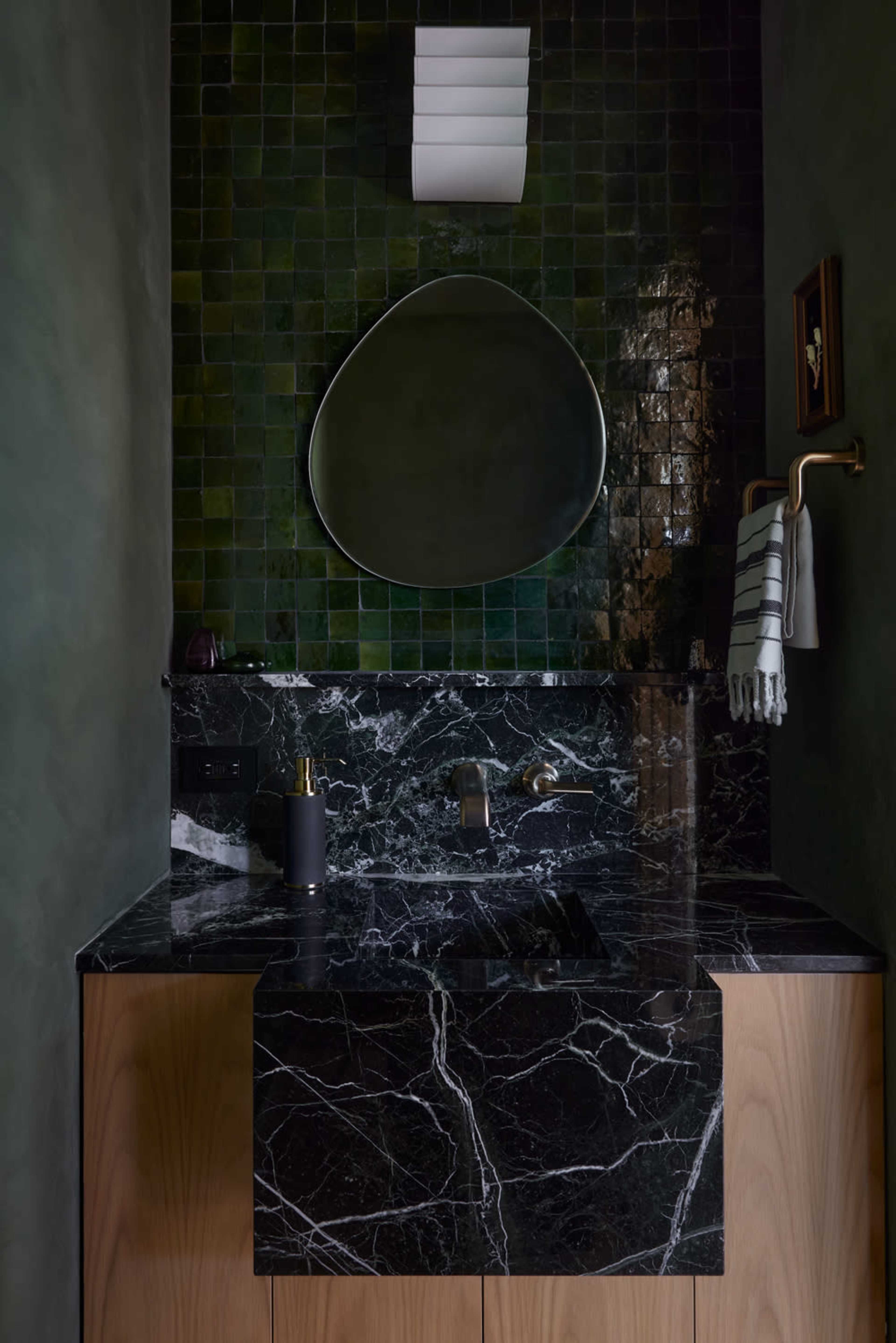 The image shows a modern bathroom with a black marble countertop, a green tiled wall, and a rounded mirror above the sink.