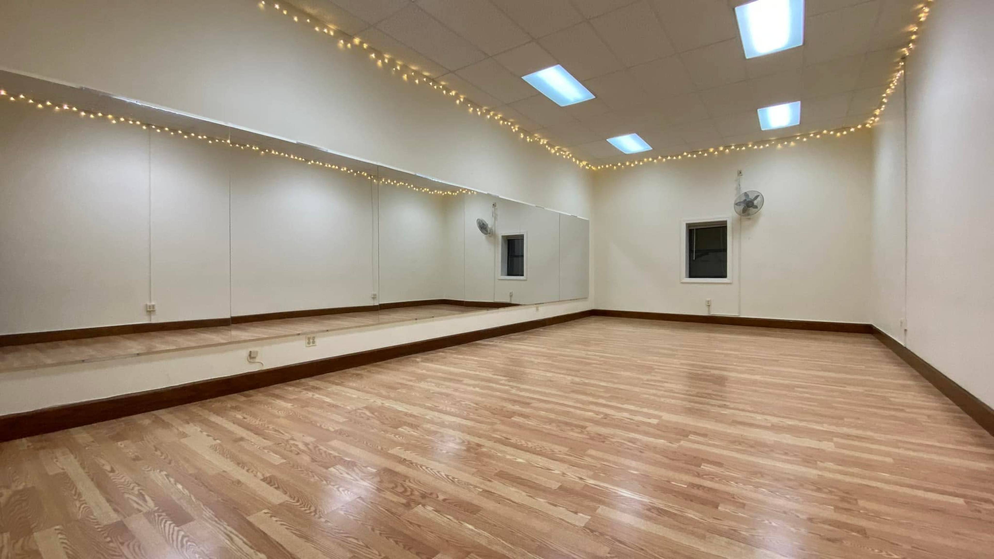 A brightly lit dance studio with wooden flooring, mirrored walls, and decorative string lights along the ceiling.