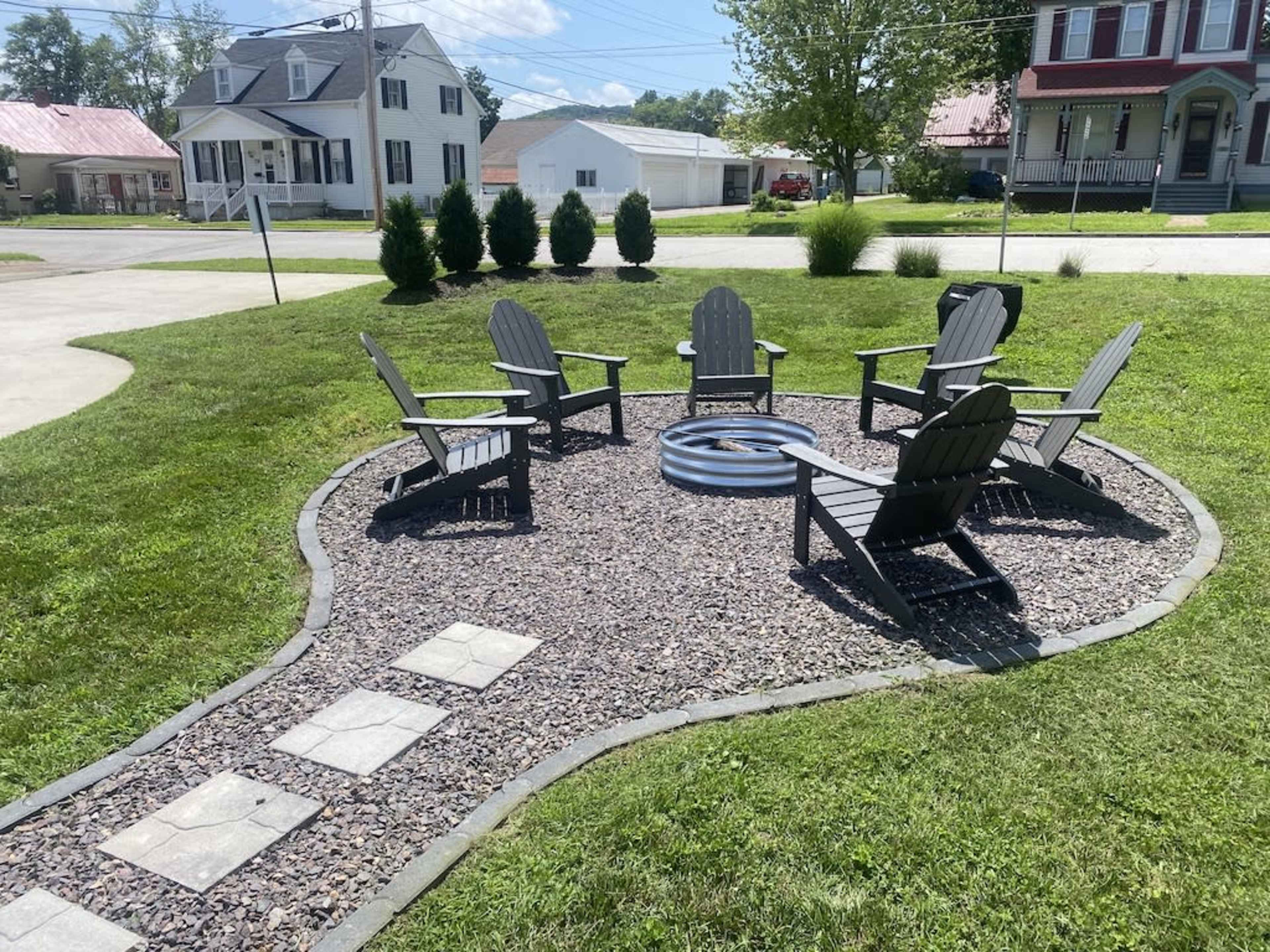 A circular fire pit area features six wooden chairs arranged around a stone circle in a grassy yard.