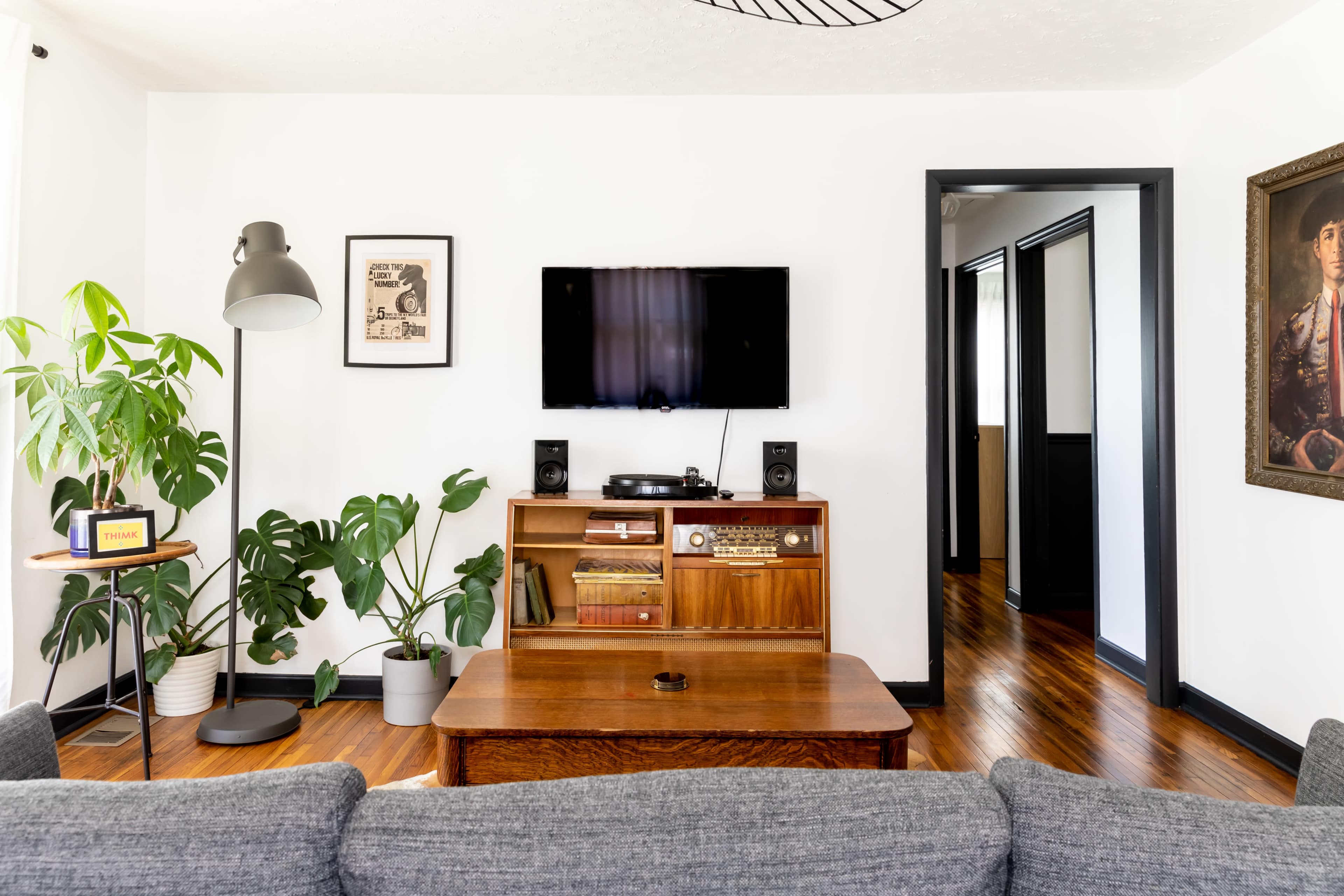 The living room features a wooden entertainment unit with a television mounted on the wall, flanked by speakers, and a coffee table in front with a large plant beside it.