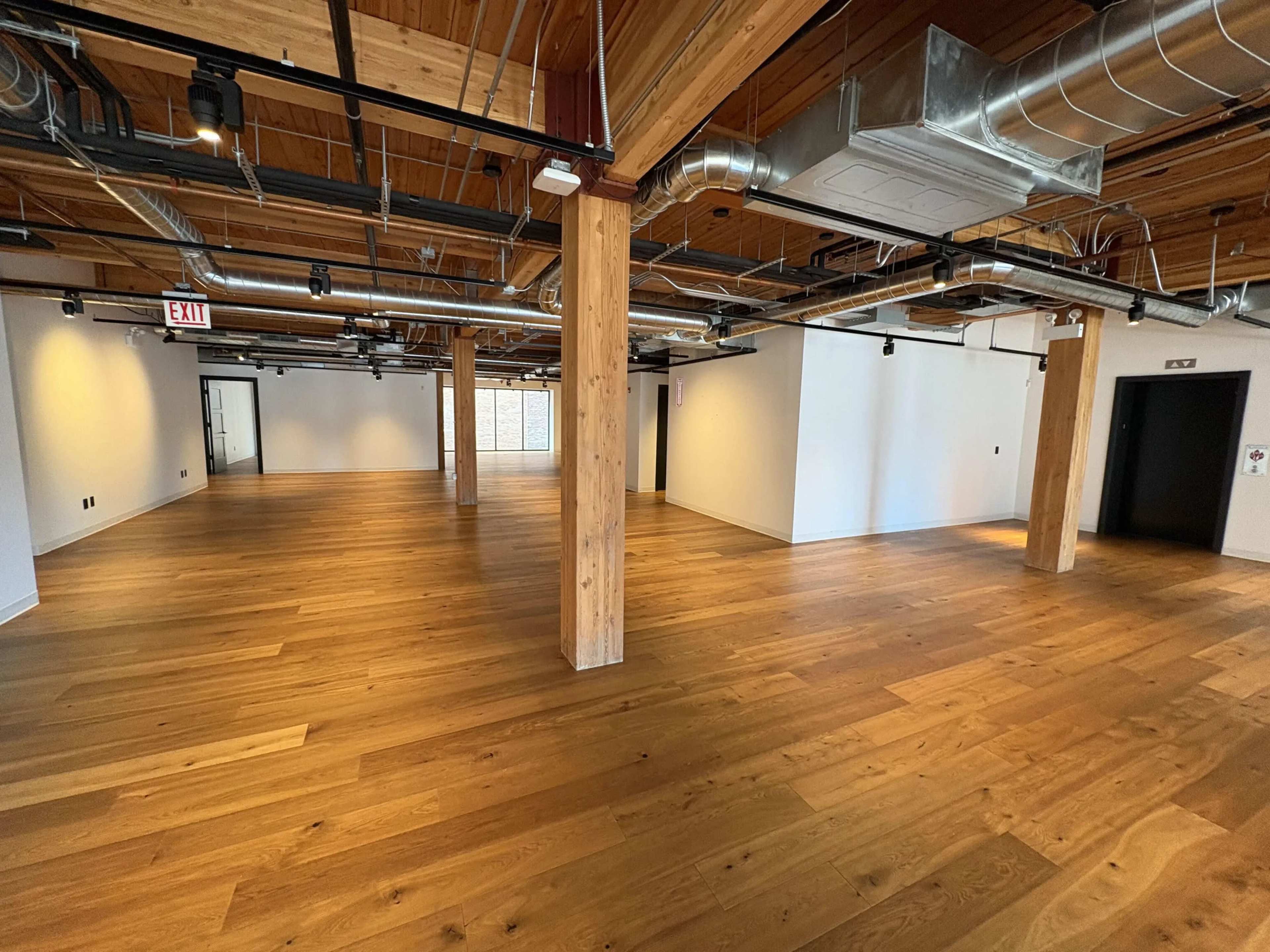 The image shows a spacious, empty office interior with wooden floors, exposed beams, and industrial piping, featuring multiple doorways and an absence of furniture.