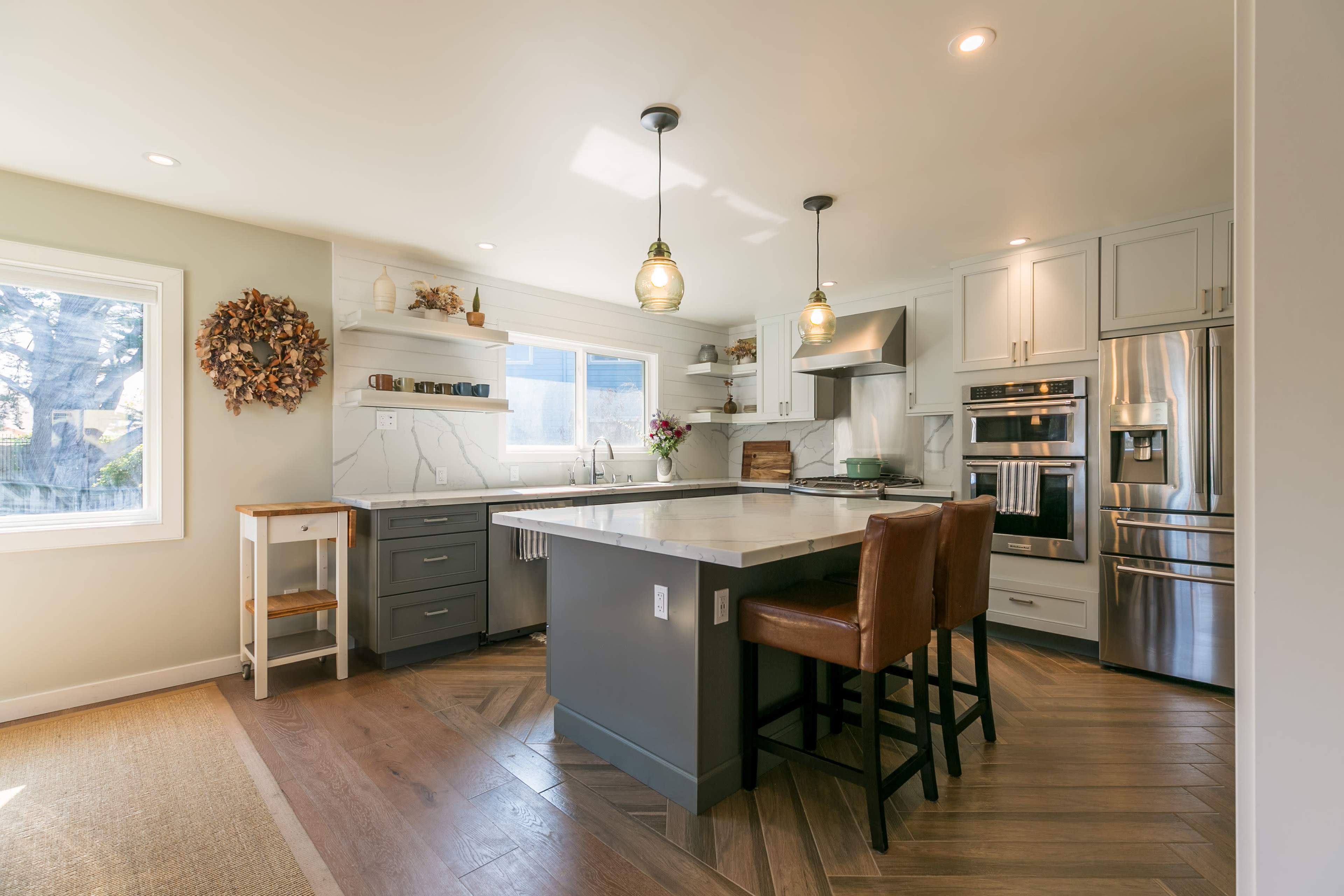 A modern kitchen features a large island with two brown barstools, stainless steel appliances, and white cabinetry, illuminated by pendant lights.