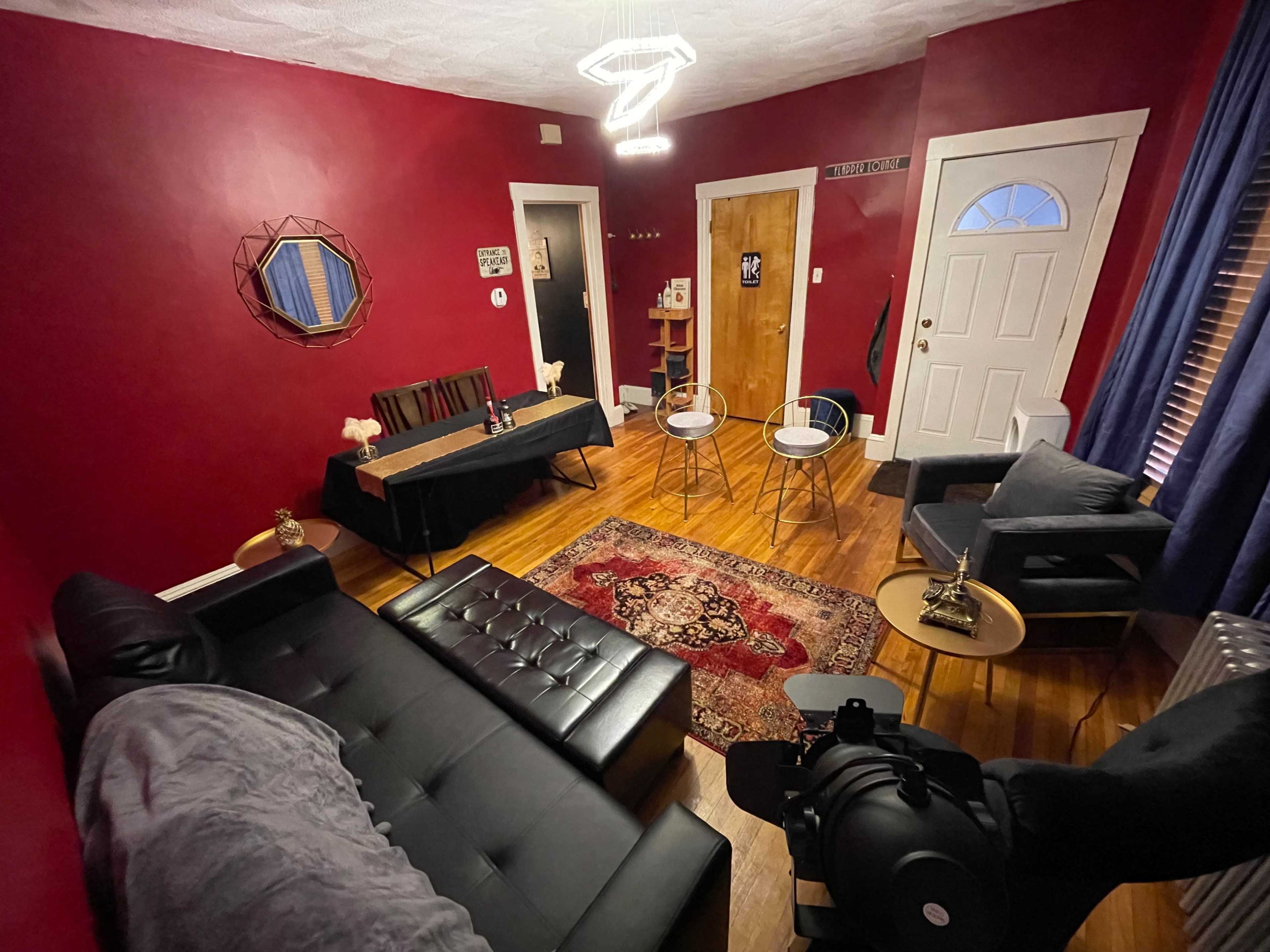 The room features a dark red wall, a black sofa, a table set for two, and a pair of chairs facing a door and a window.
