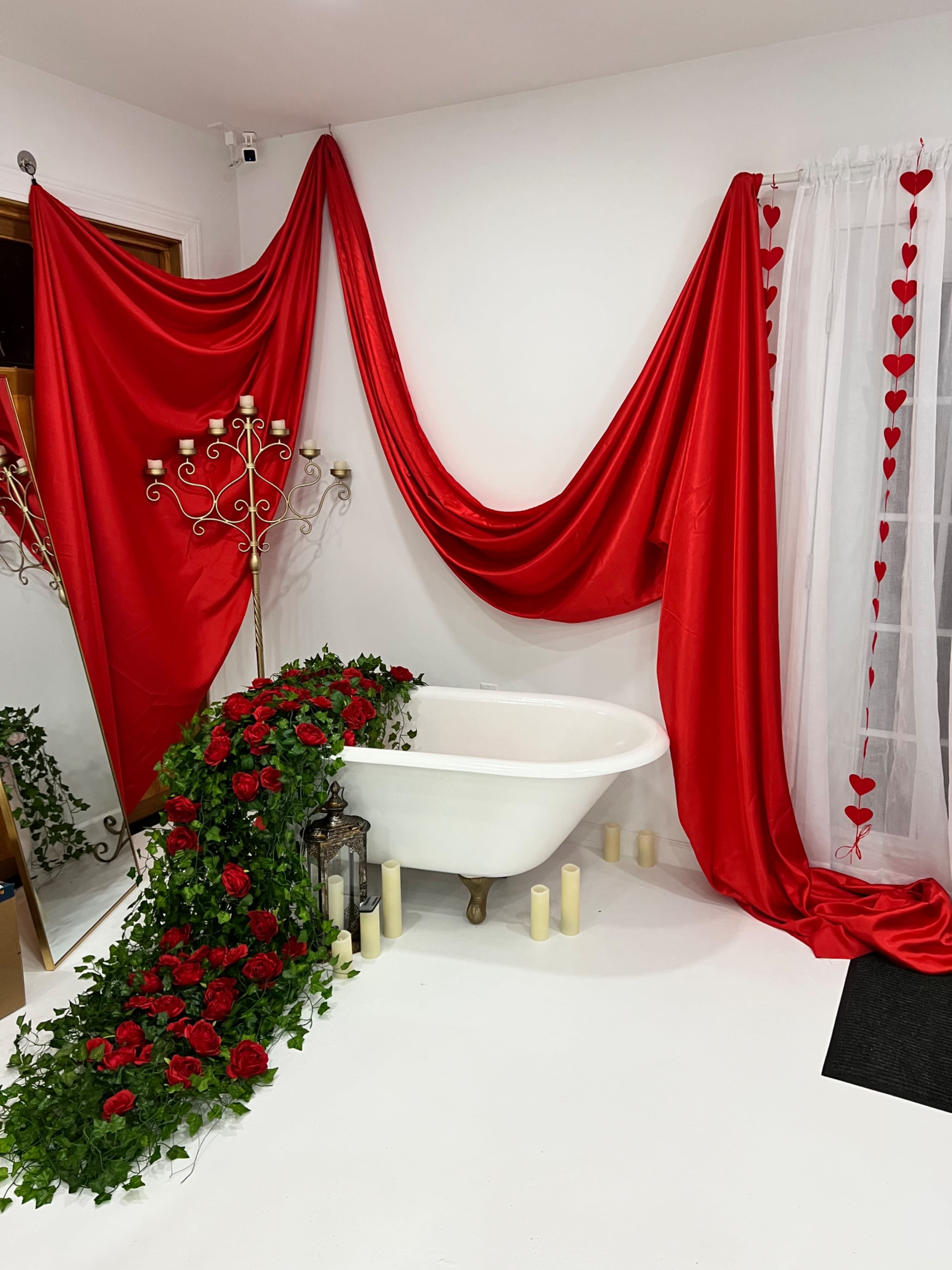 A white bathtub is adorned with red fabric and greenery, surrounded by candles and a decorative lantern.