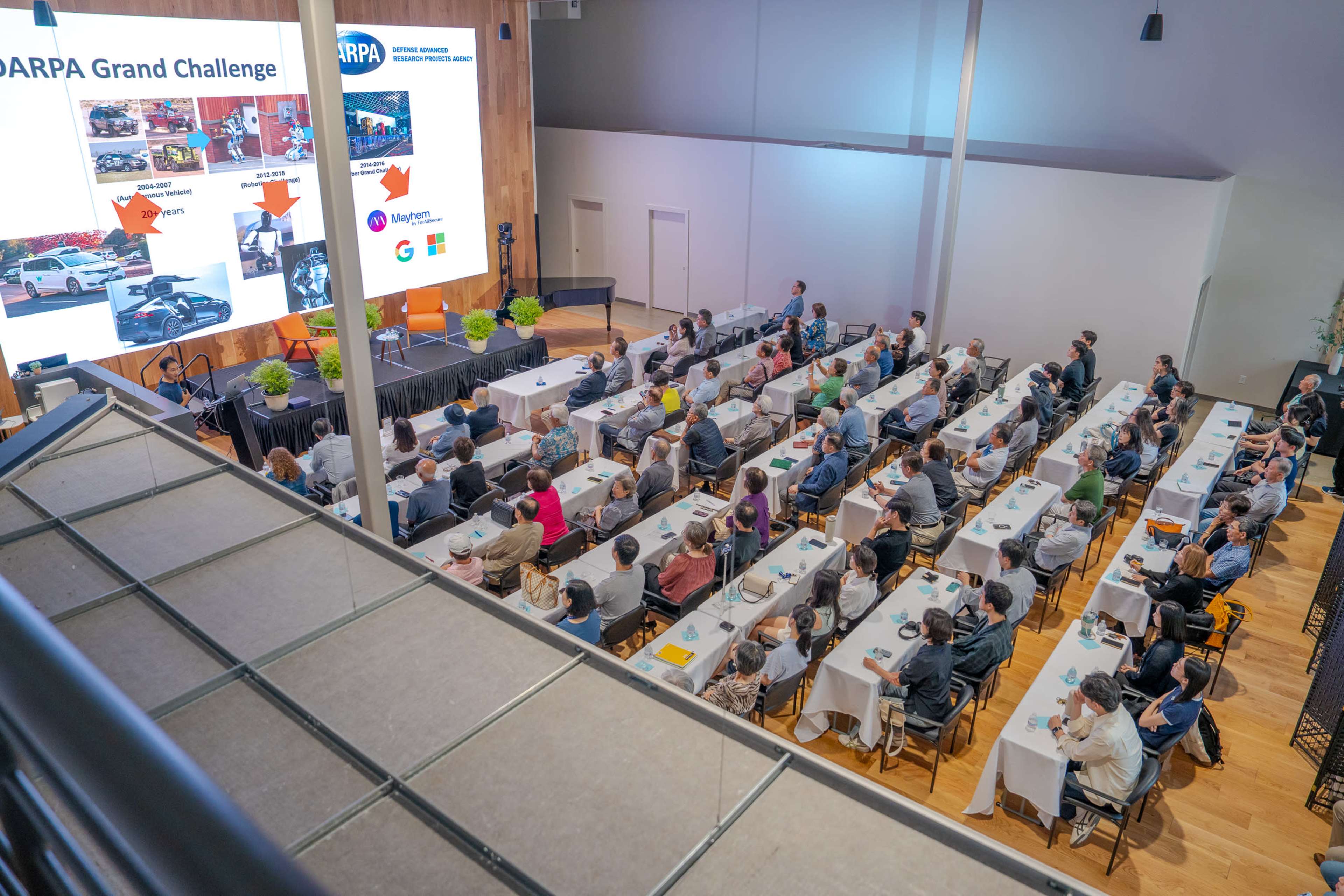The image shows a large audience seated in a conference room with tables, watching a presentation on a screen displaying the DARPA Grand Challenge.