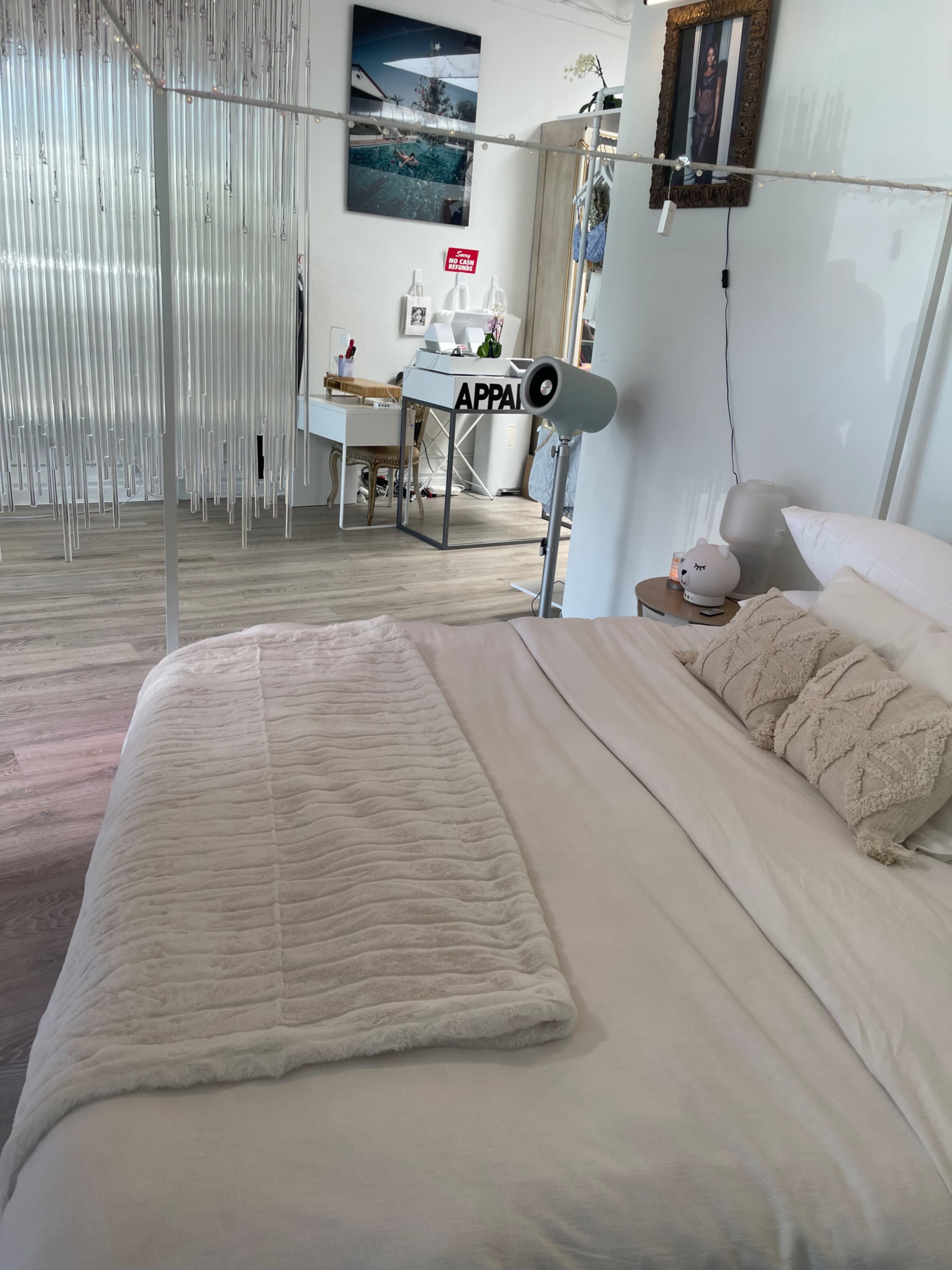 The image shows a neatly arranged bedroom with a bed in the foreground, a desk and chair in the background, and decor including a framed picture on the wall.