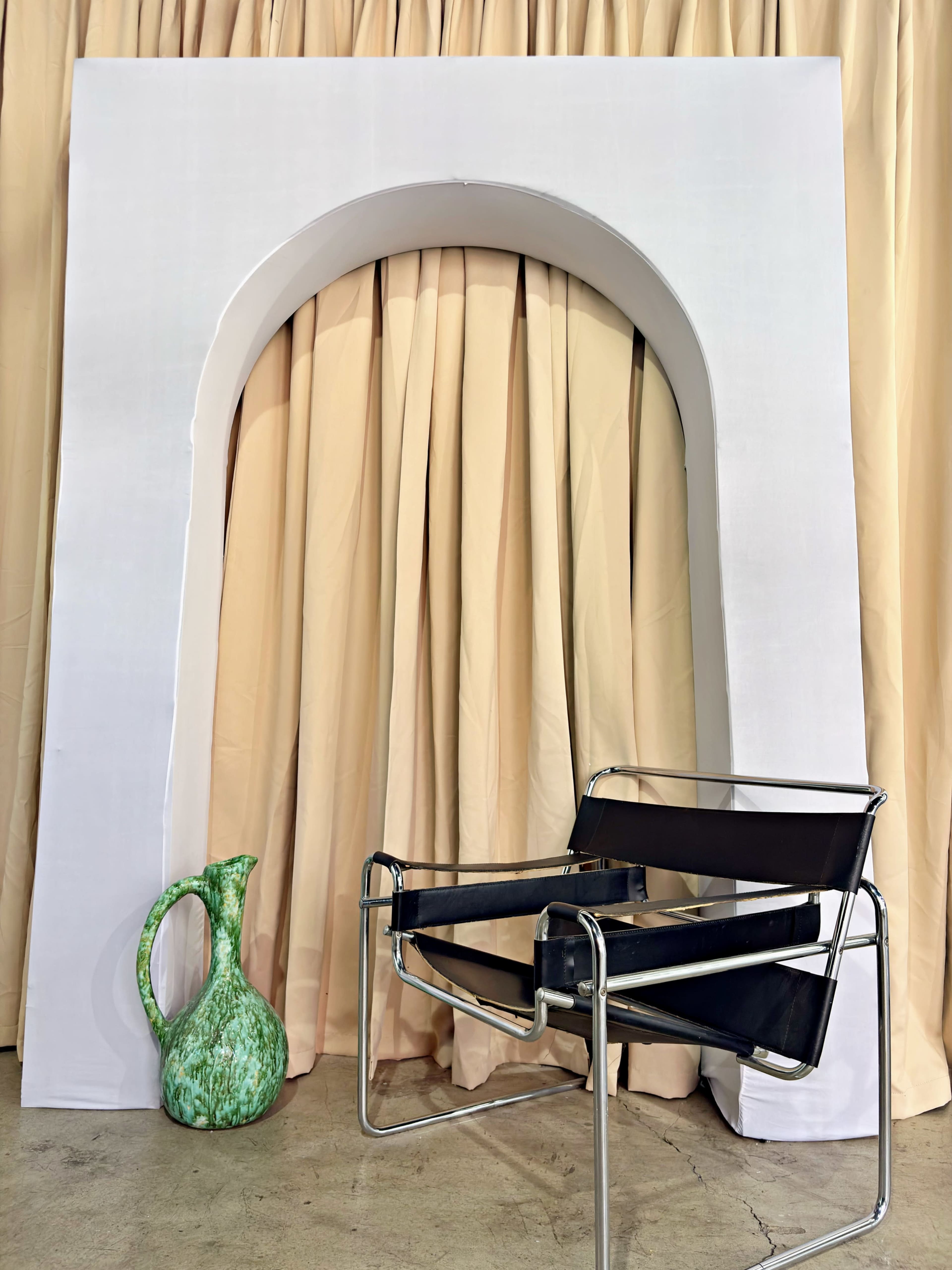 The image shows a minimalist setup featuring a large white arch structure, a black chair, and a green decorative pitcher next to it, against a backdrop of beige curtains.