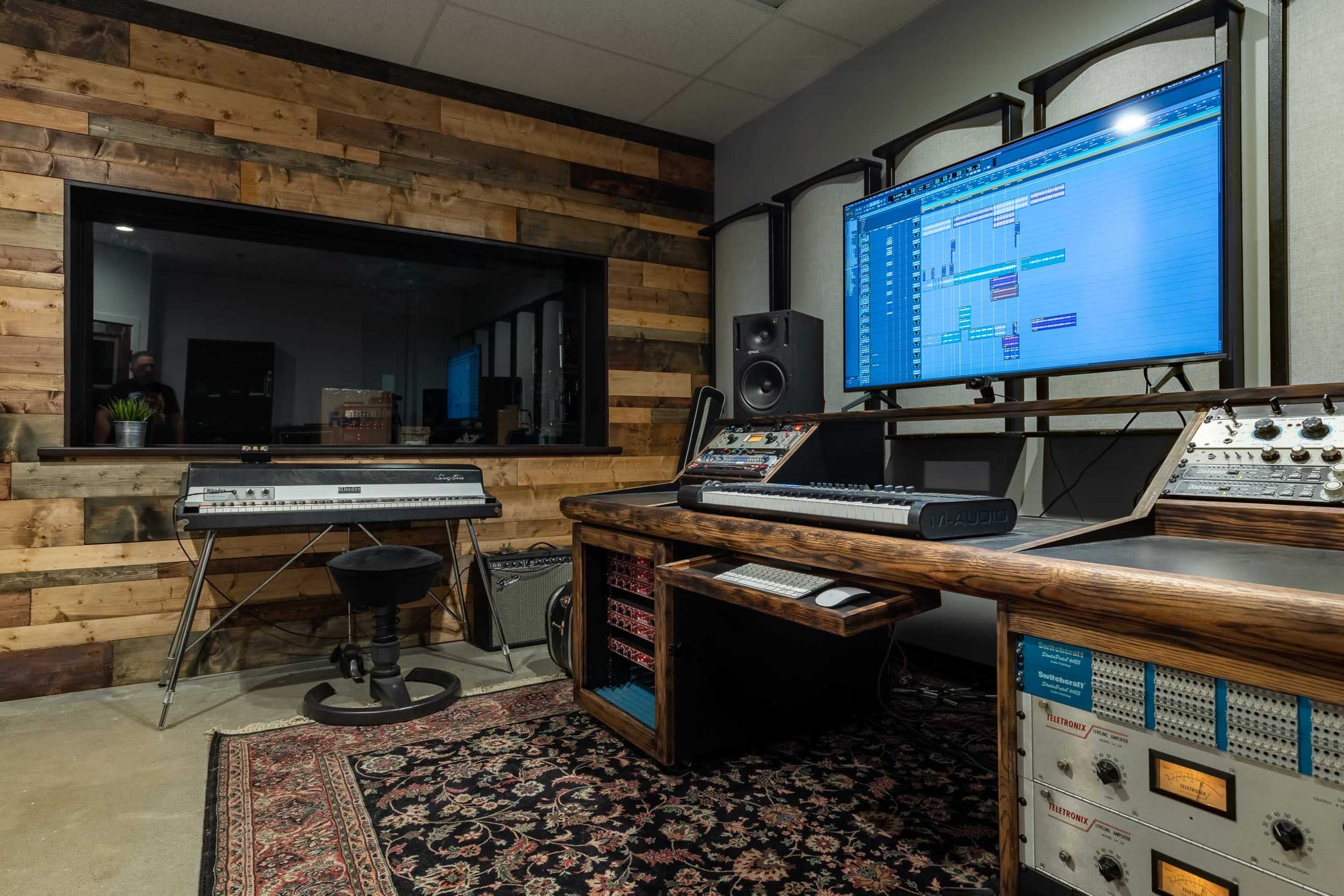 A music studio with a wooden wall, a computer screen displaying digital audio tracks, a keyboard, and various audio equipment.