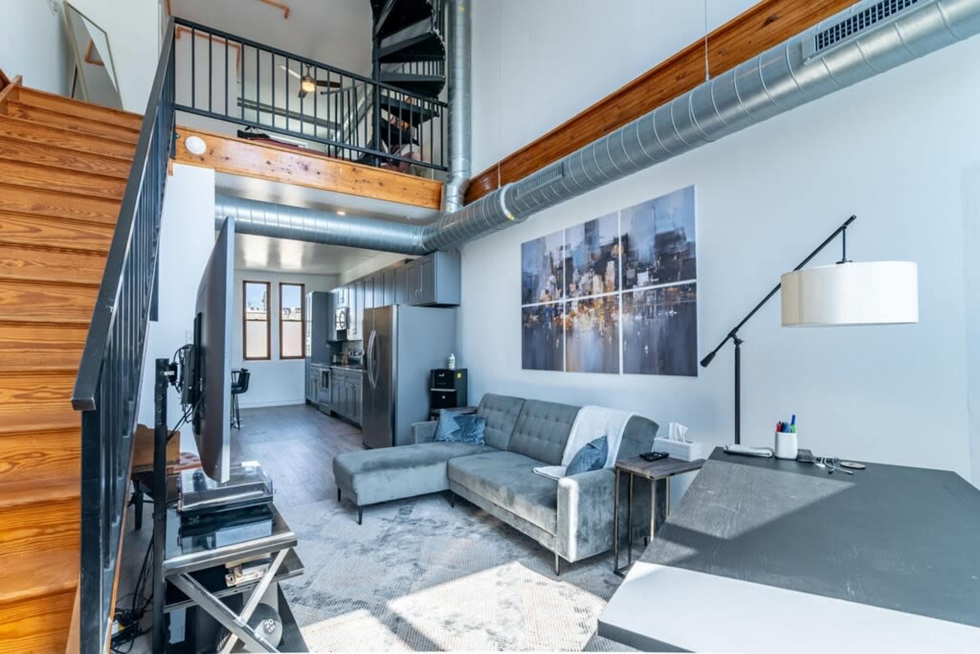 The image shows a modern, open-concept living area with a staircase, kitchen, and living room featuring a gray sofa and a large wall art piece.