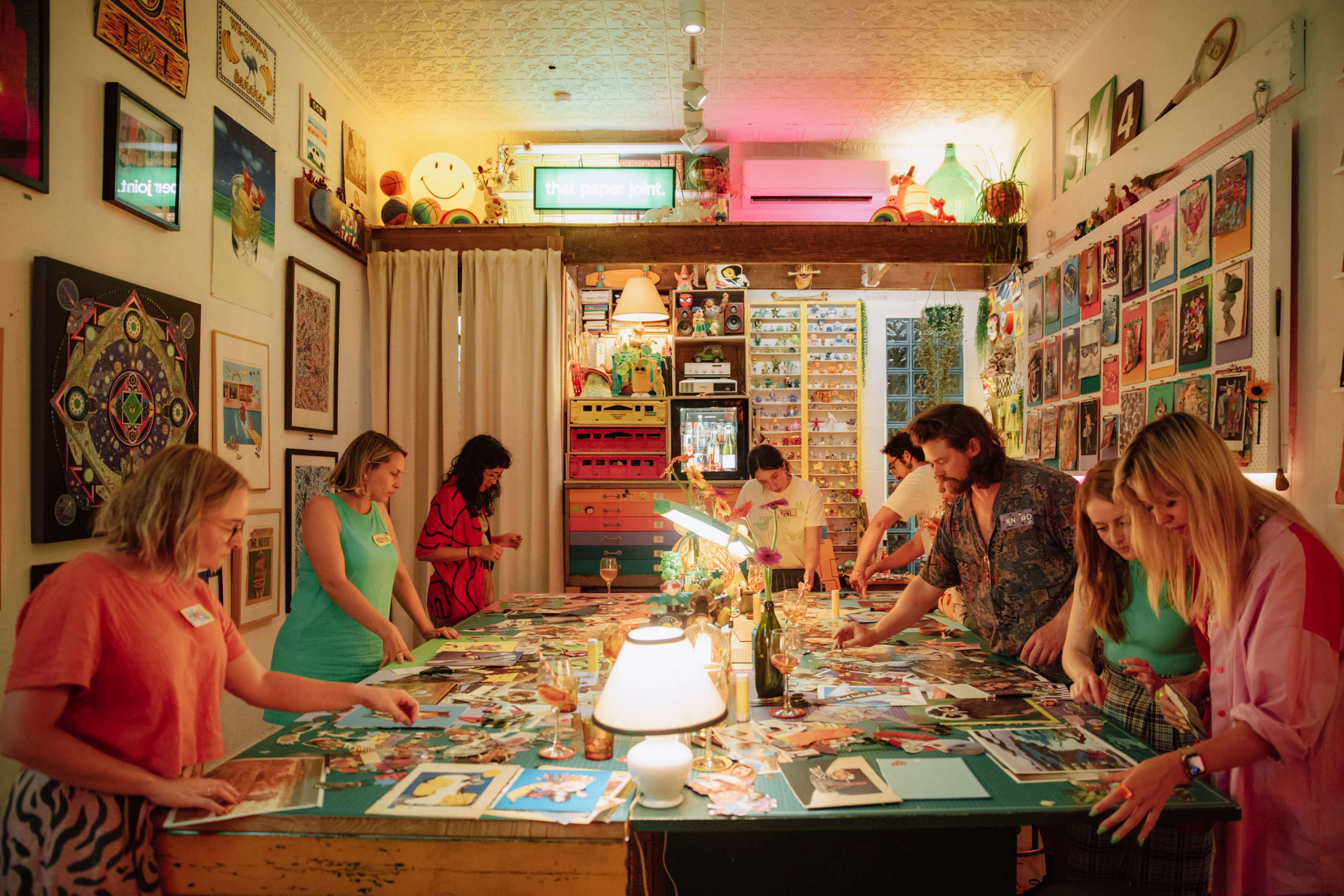 A group of people sorts through photographs at a brightly lit creative workspace filled with various artworks and decorative items.