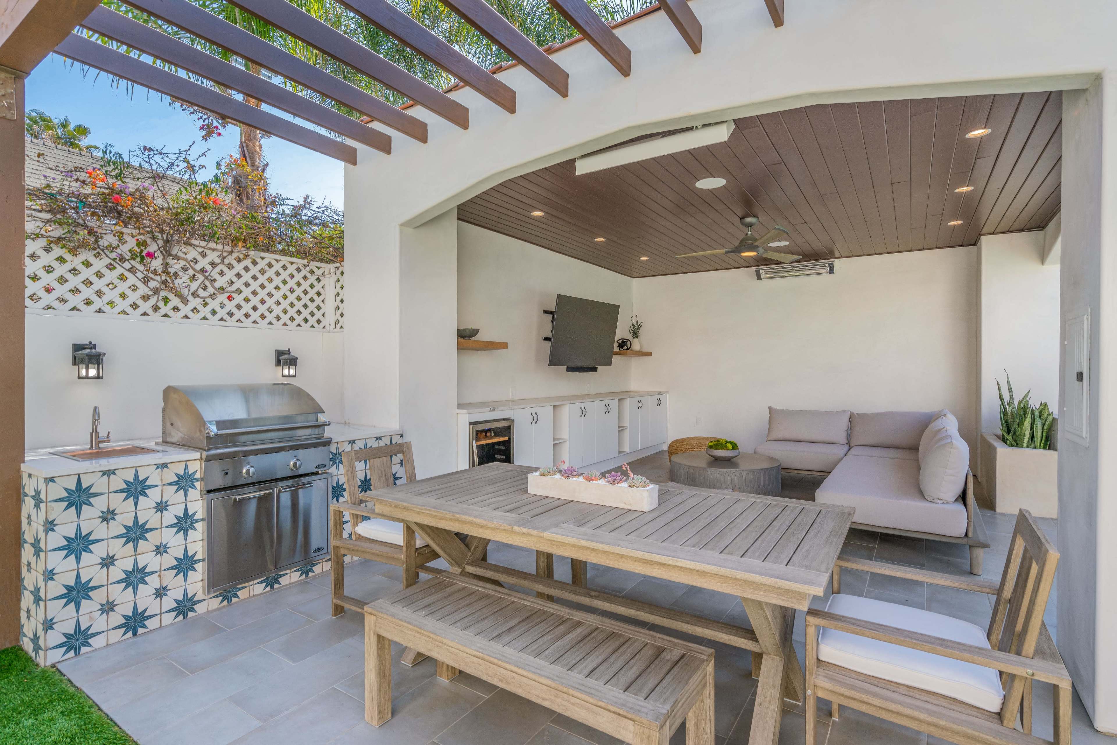 The image shows a modern outdoor kitchen and dining area with a barbecue grill, seating, and a flat-screen TV under a shaded pergola.