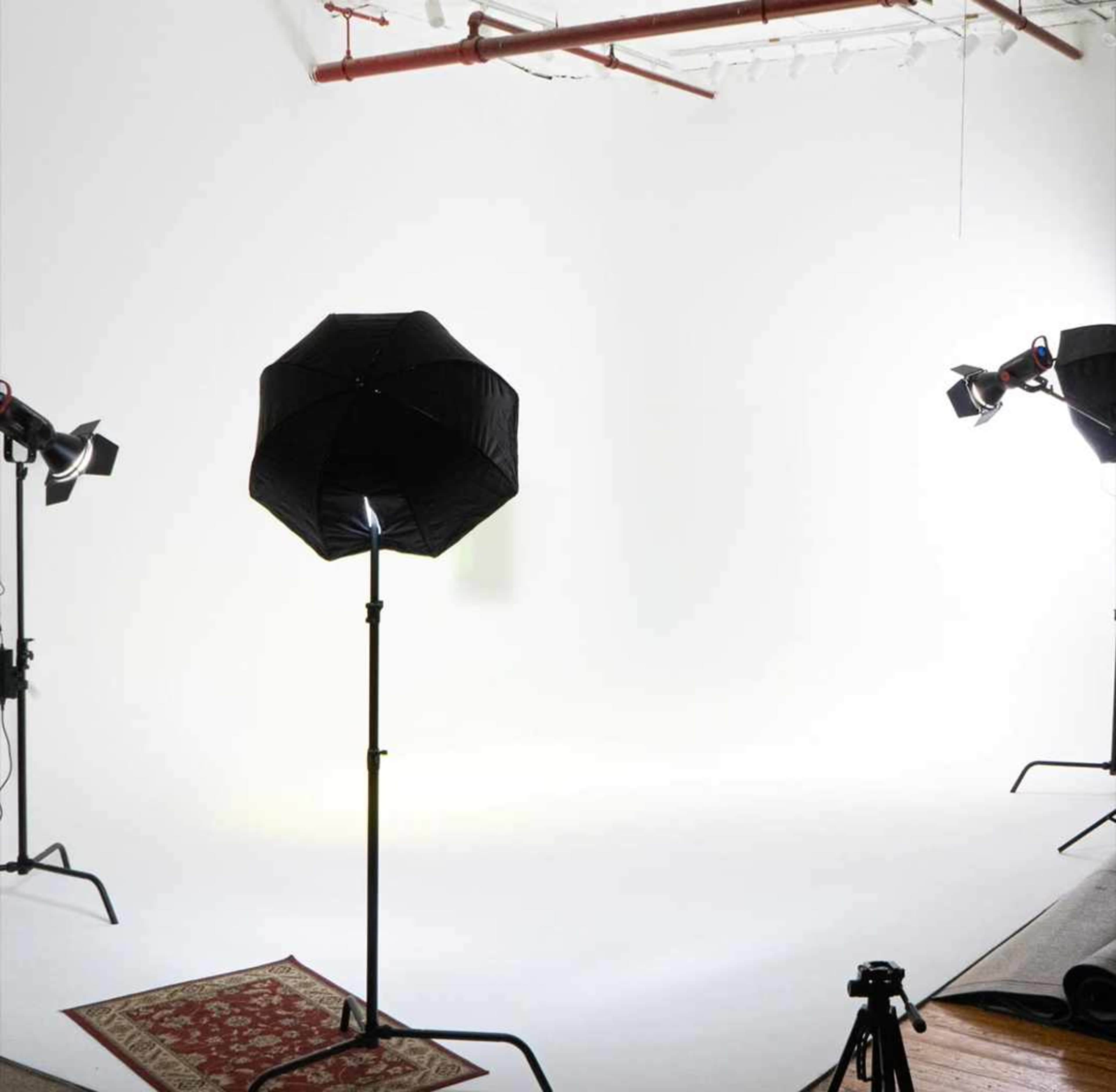 A photography studio setup with light stands, softboxes, and a patterned rug on a plain white backdrop.