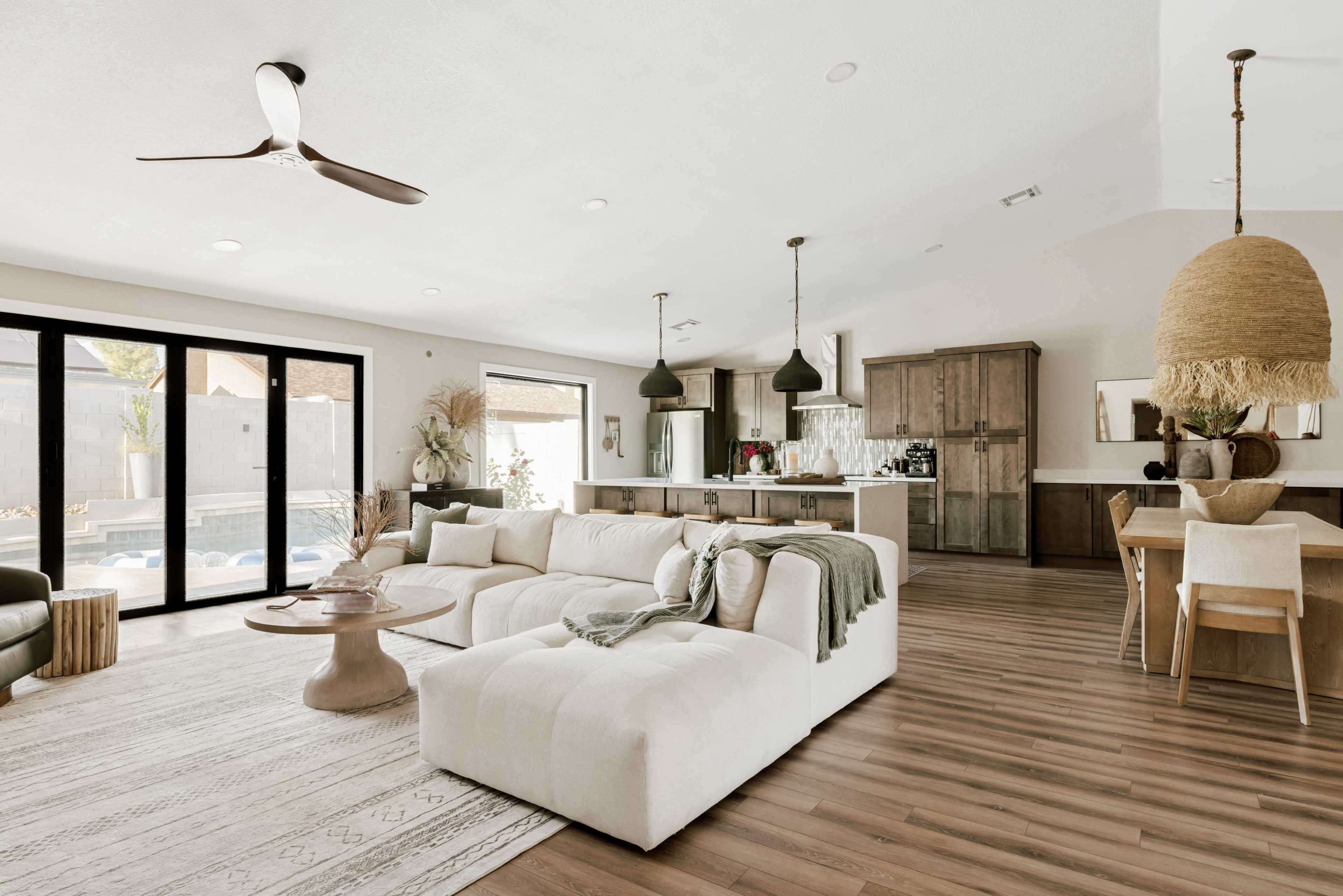 The spacious living area features a large sectional sofa, a dining table, and an open kitchen, all illuminated by natural light from large sliding glass doors.