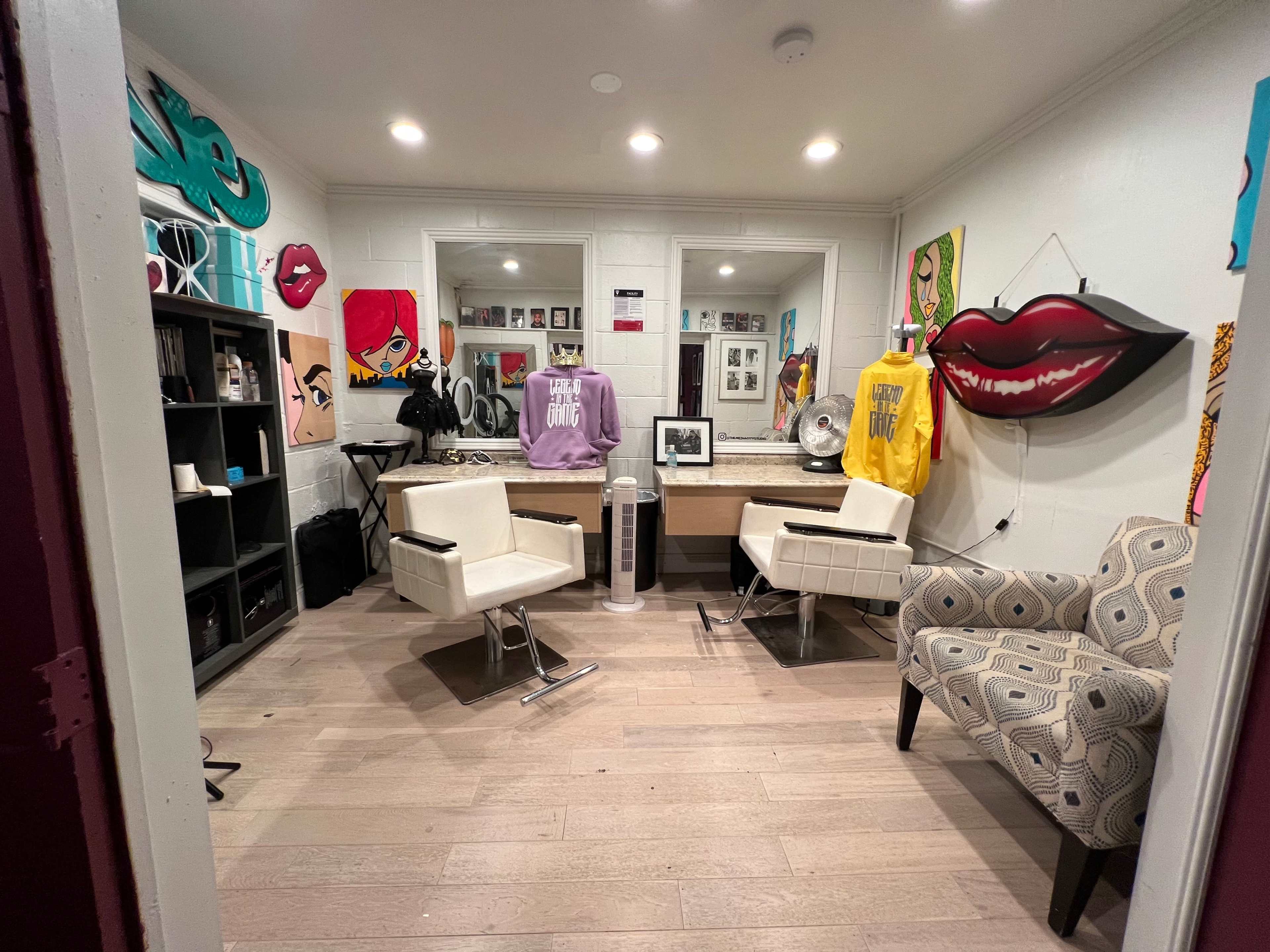 A stylized salon interior featuring two white chairs, a large vanity mirror, and various colorful artworks on the walls.