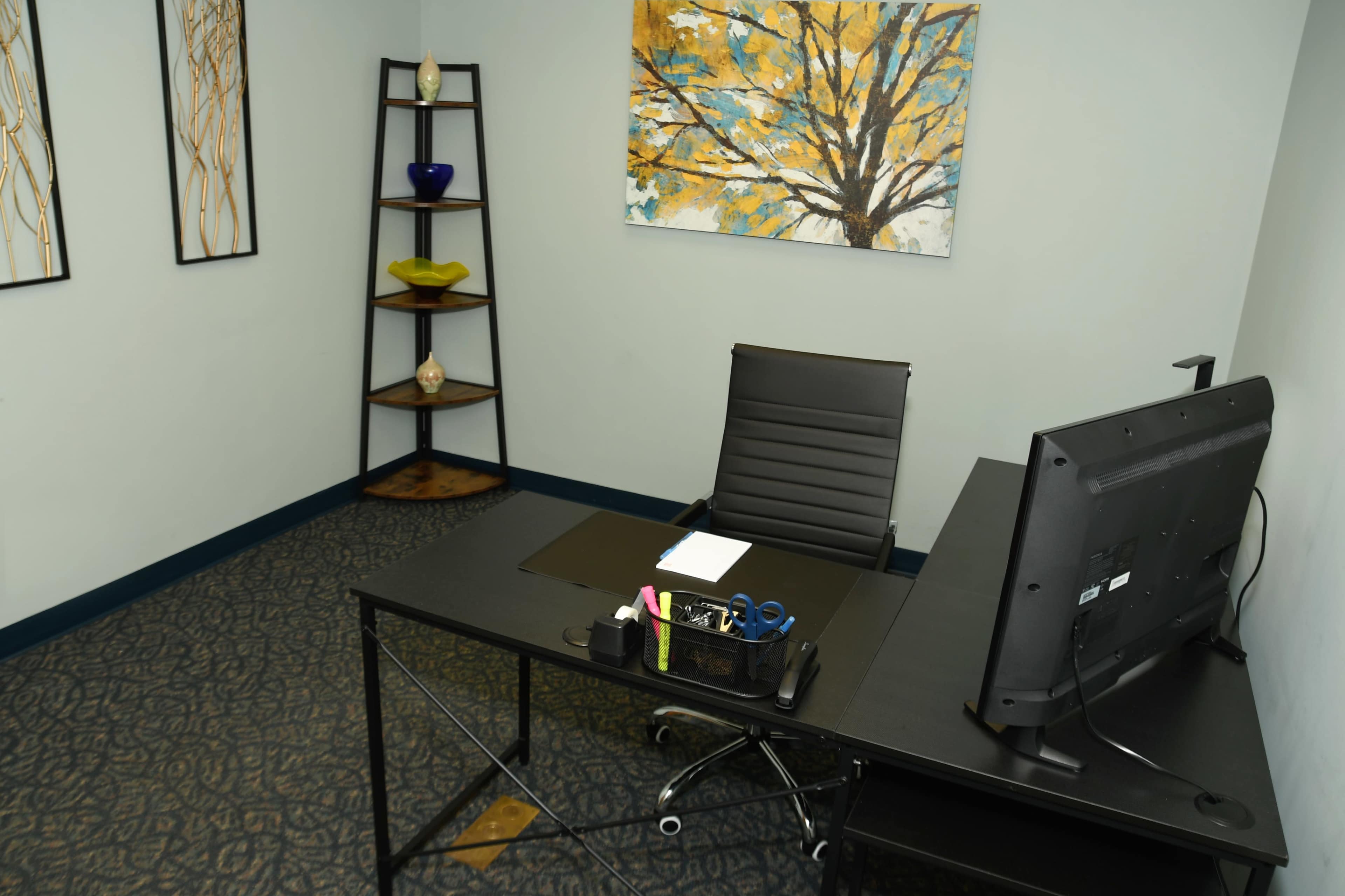 The image shows a minimalist office space featuring a black desk with a monitor, a chair, and a tiered shelf displaying decorative items and artwork on the wall.
