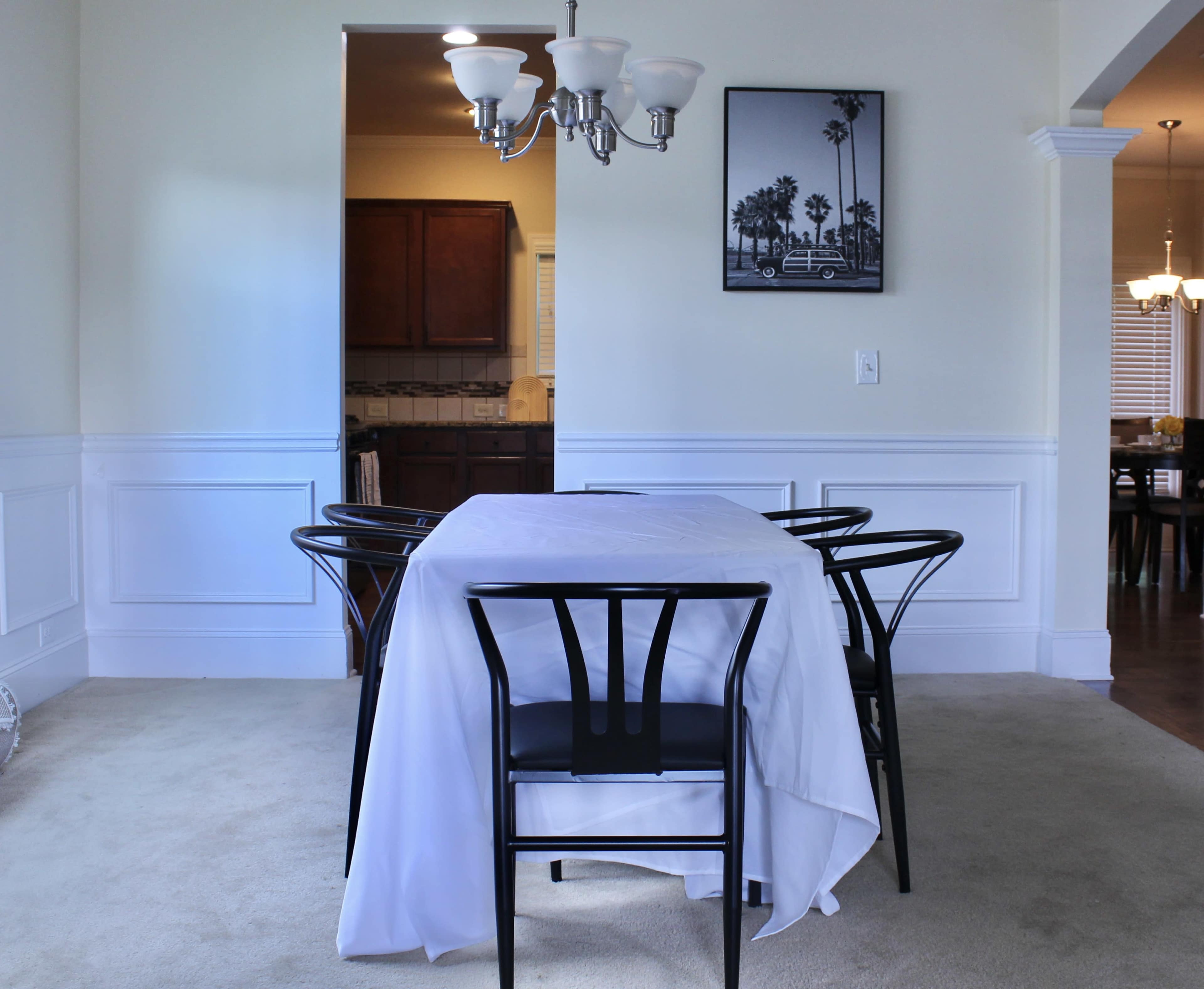 A dining area features a table covered with a white tablecloth and black chairs, with a chandelier overhead and a framed photo on the wall.