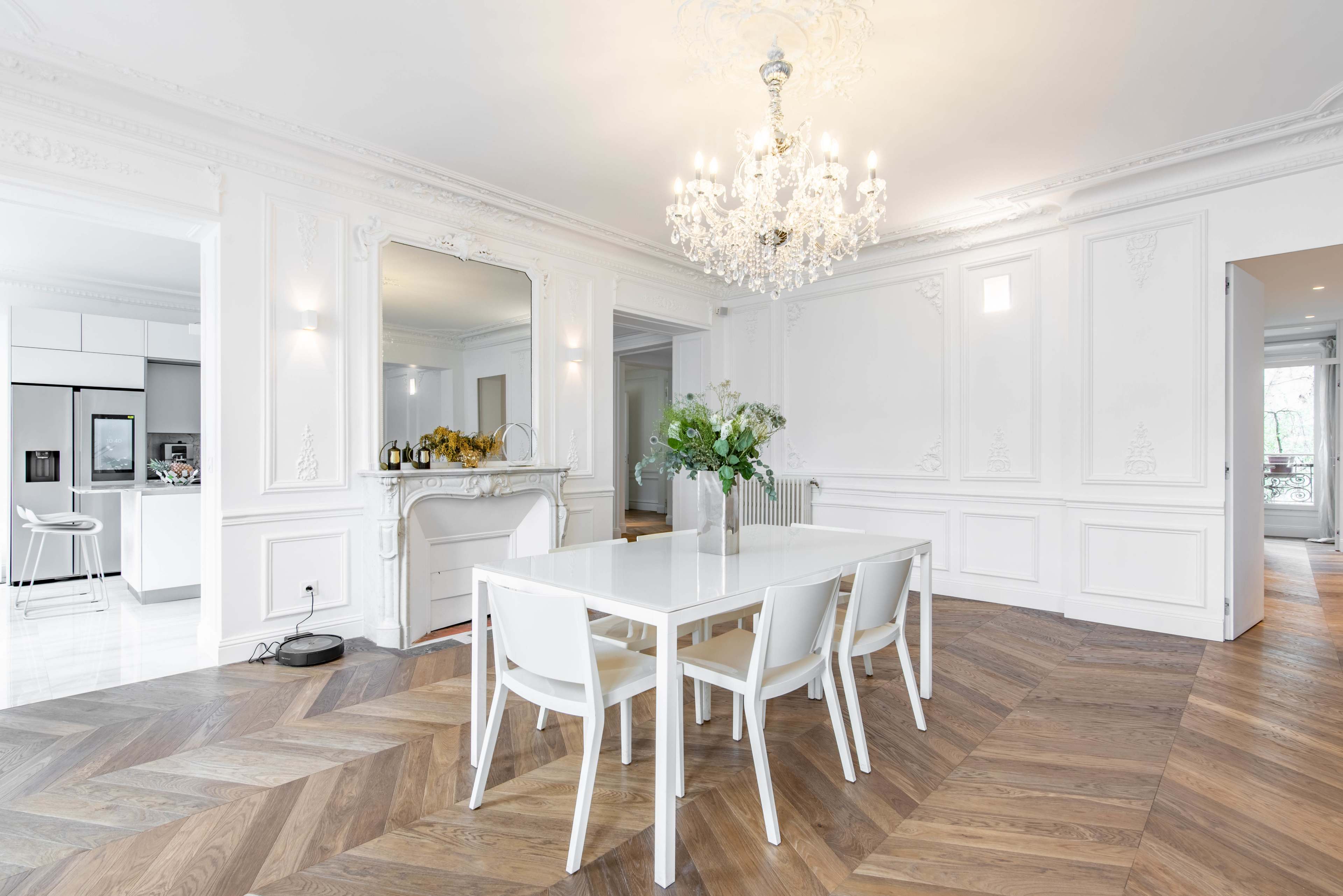 The image depicts a modern dining room featuring a large white table surrounded by white chairs, a chandelier overhead, and elegant wall paneling with a patterned wooden floor.
