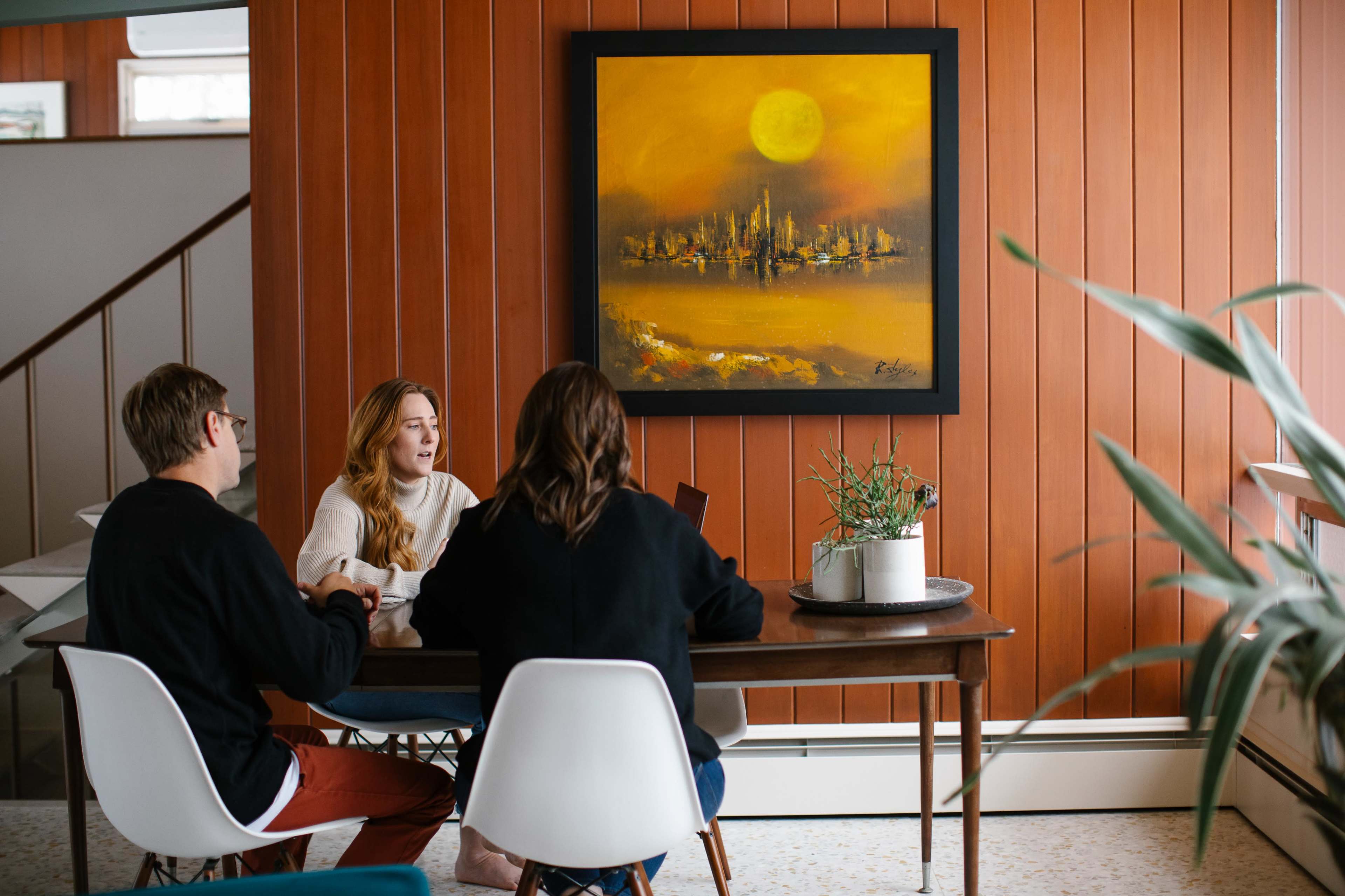 Three people are engaged in conversation at a dining table beneath a large, abstract painting on a wall with wooden paneling.