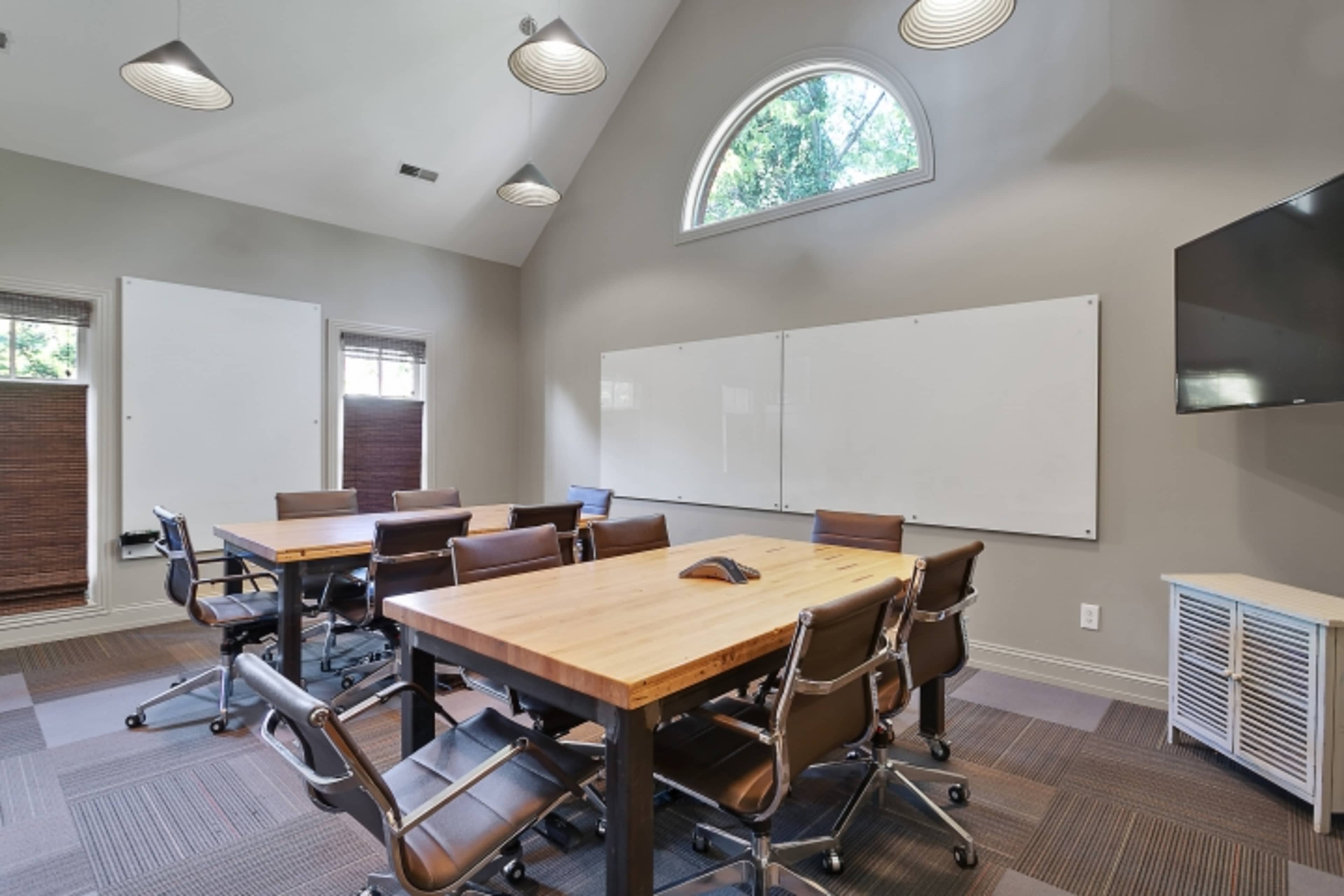 A modern conference room features multiple wooden tables, ergonomic chairs, whiteboards, and a wall-mounted television.