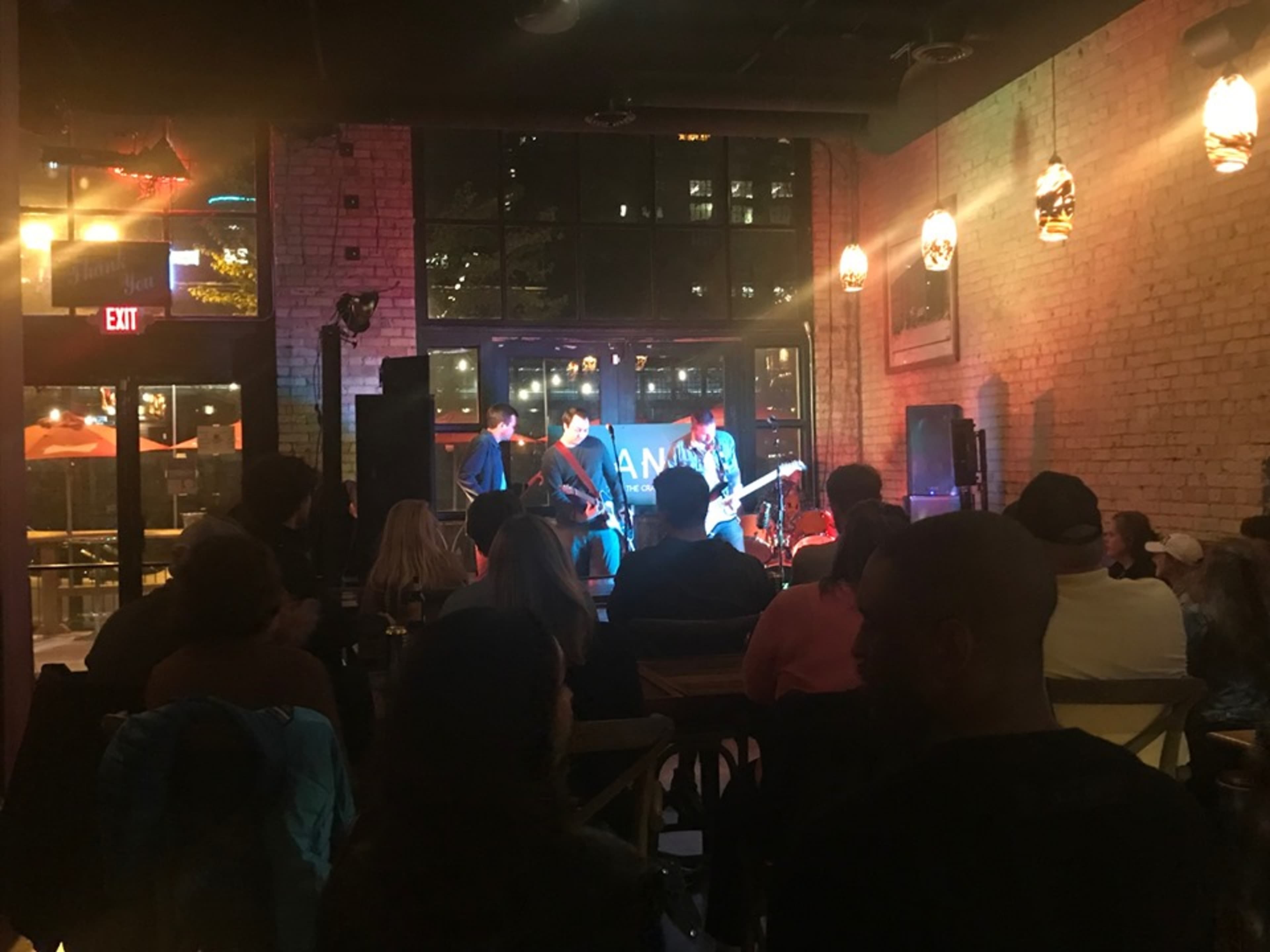 A small crowd watches a live band perform inside a dimly lit venue with large windows.