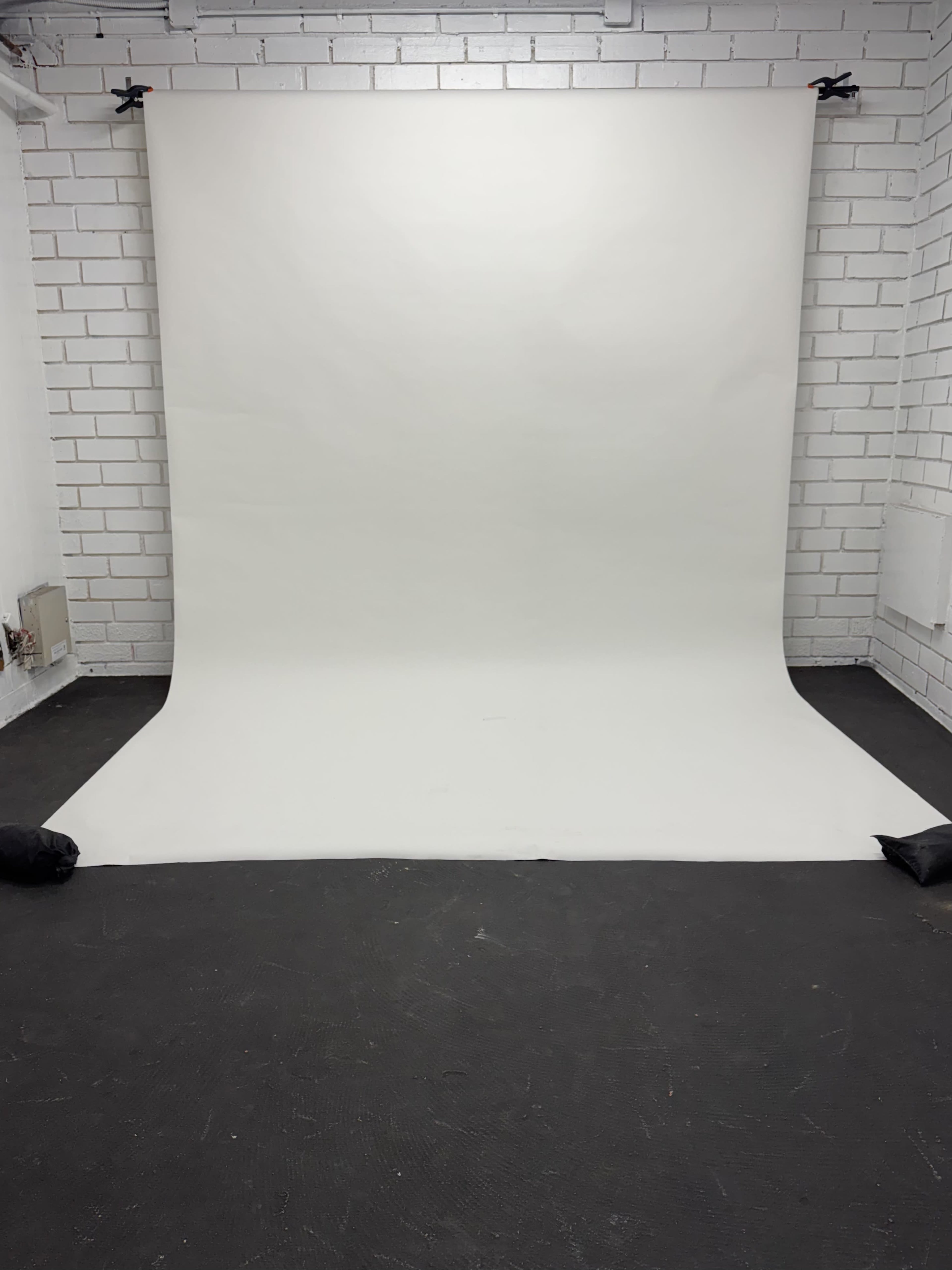 A photography studio features a large white backdrop with black weights holding down the corners, set against a white brick wall.