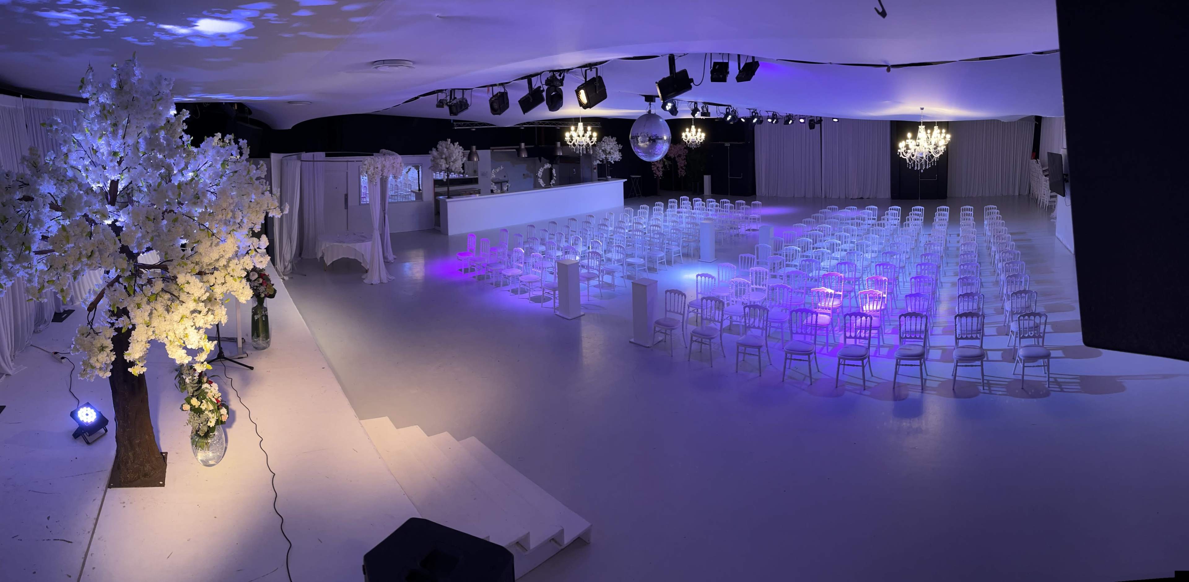 A spacious event venue is set up with rows of white chairs, illuminated by ambient lighting and decorated with artificial cherry blossom trees.