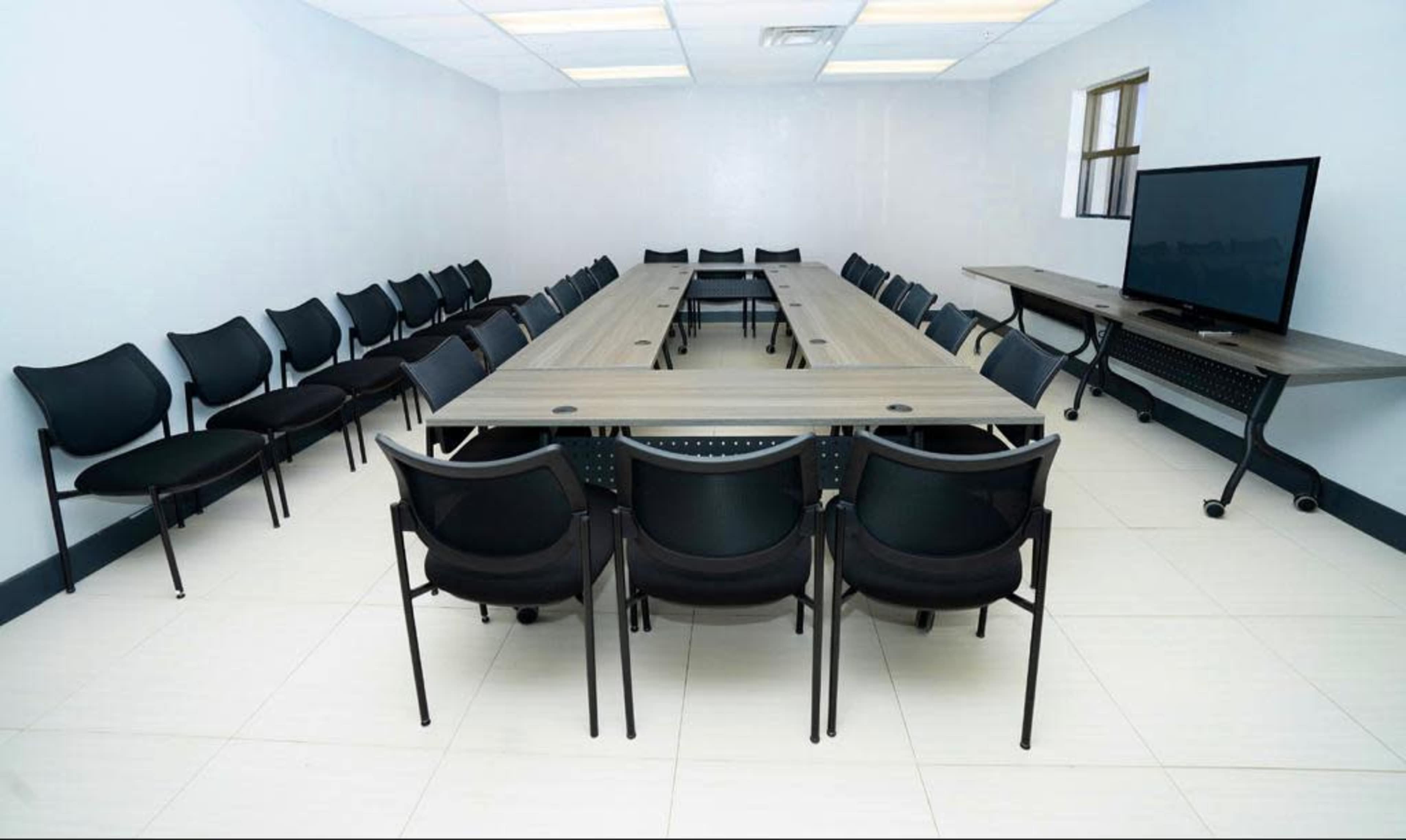 A rectangular conference room with a long table surrounded by black chairs and a television mounted on a stand against one wall.