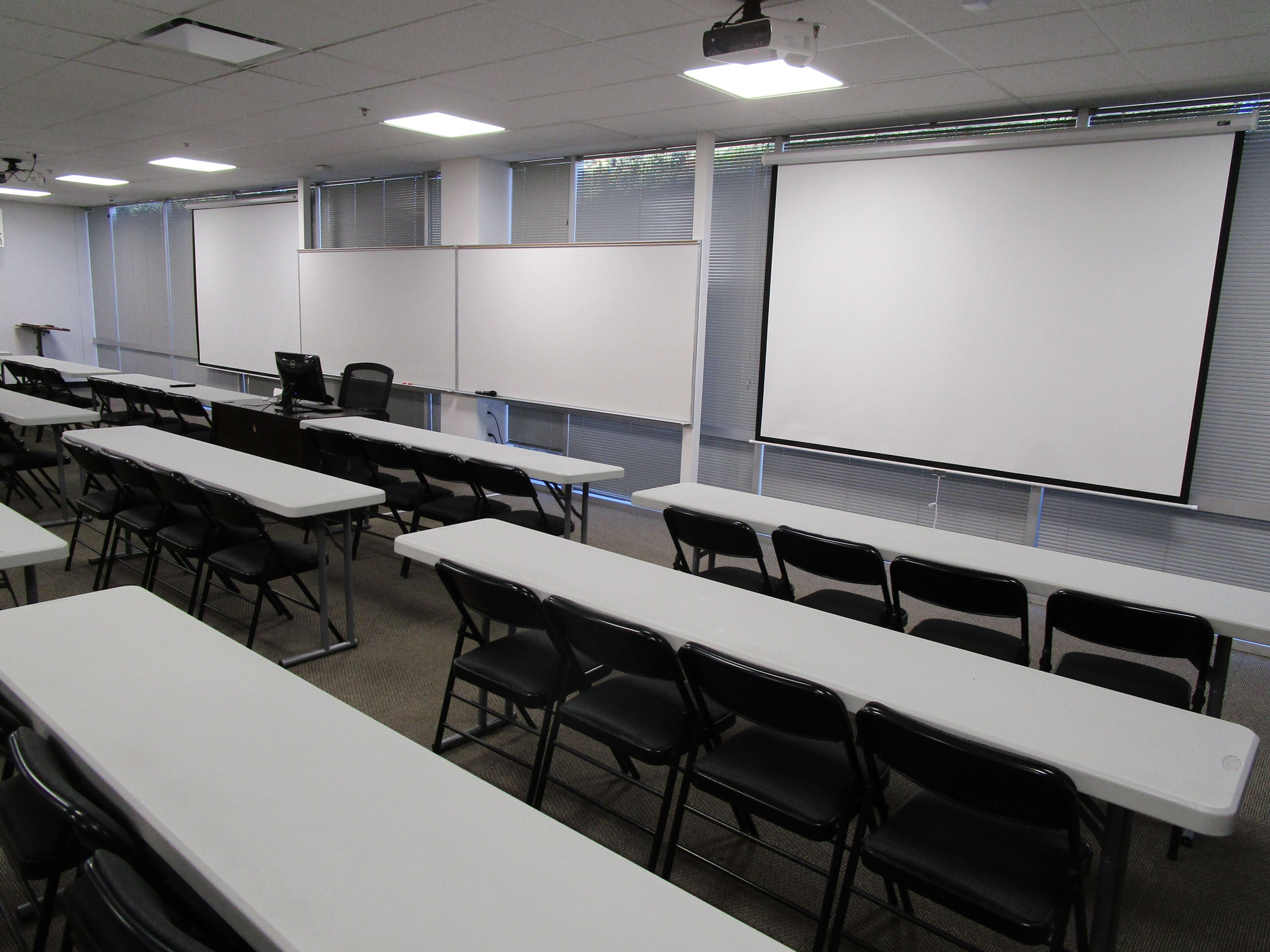A classroom setup with rows of tables and chairs arranged in front of two projection screens and whiteboards.