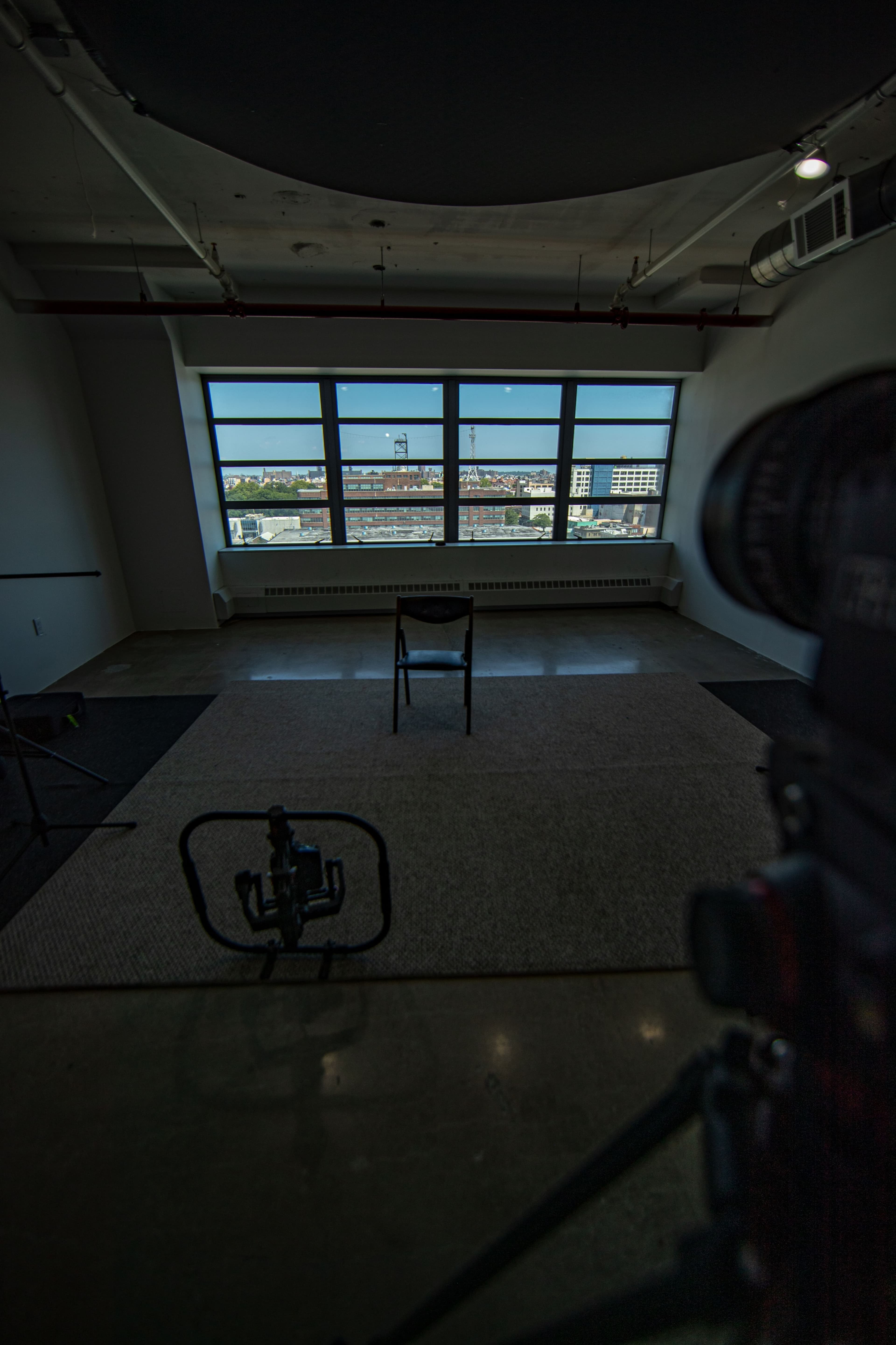 The image shows an empty room with a single chair placed in the center and large windows revealing a view of a cityscape outside.