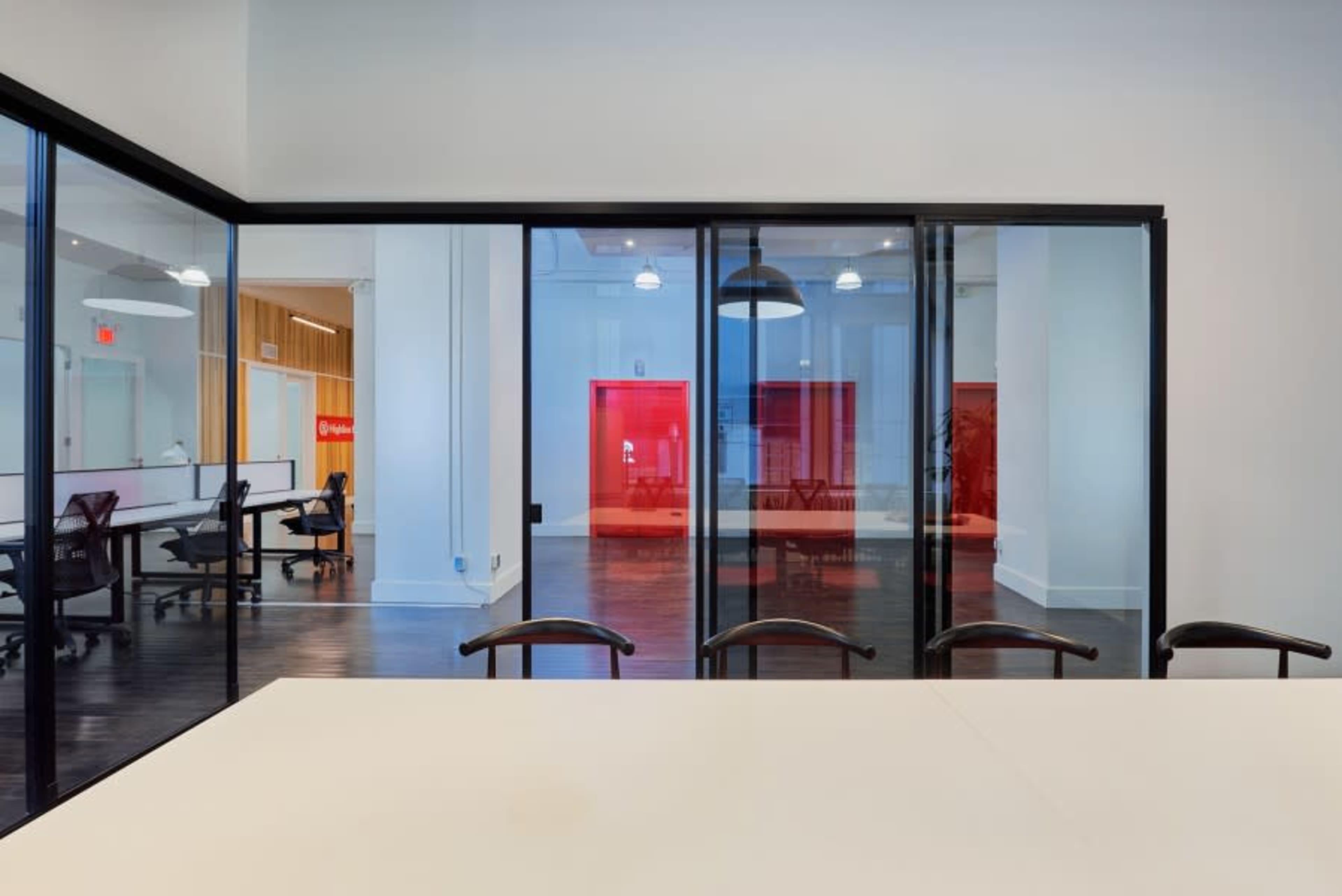 A modern office space features a conference room with a large white table and glass walls, overlooking an area with red-lit doors and empty desks.
