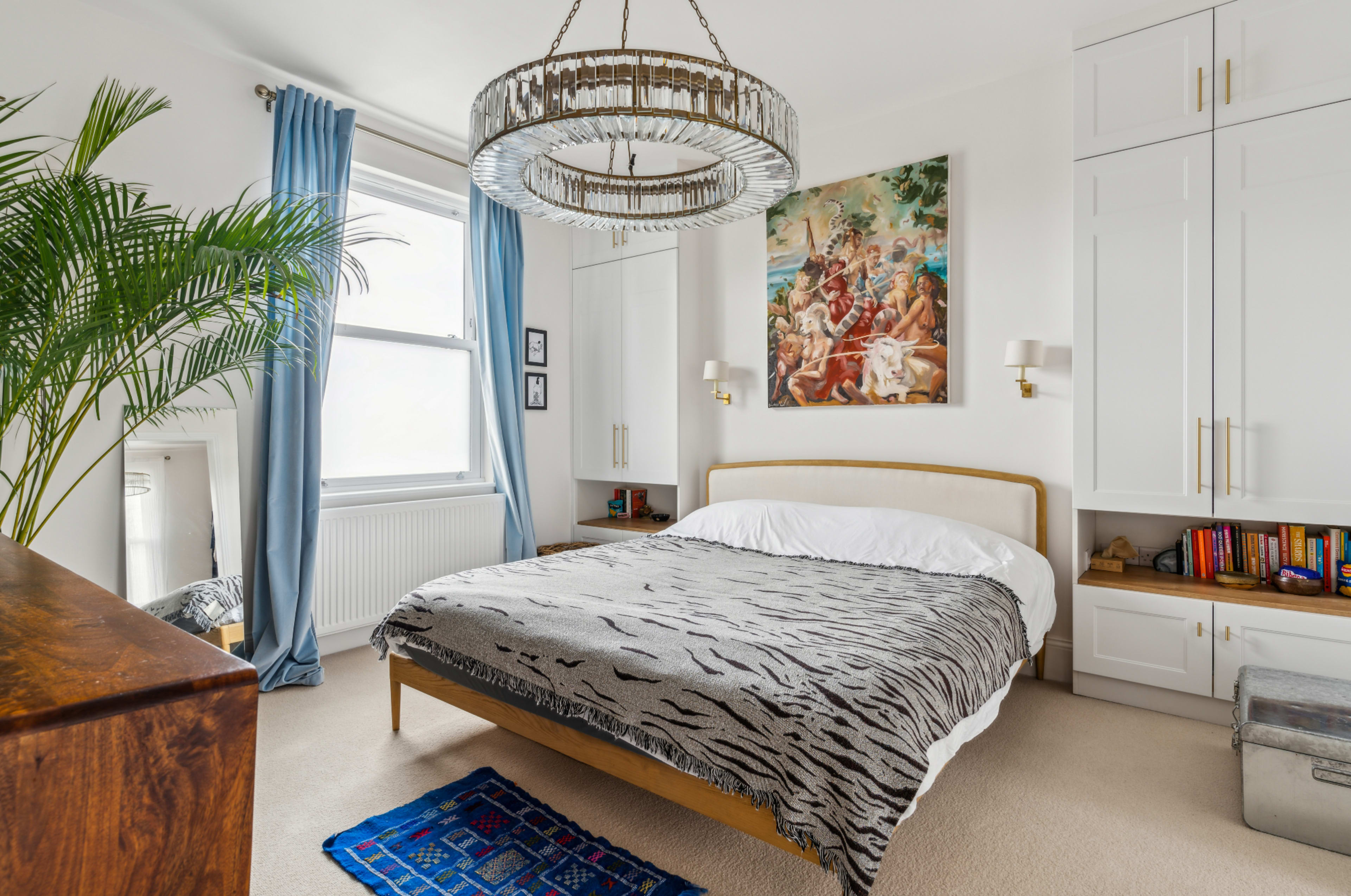 A neatly arranged bedroom with a bed covered in a zebra-striped blanket, a large chandelier overhead, and a colorful artwork hanging on the wall.
