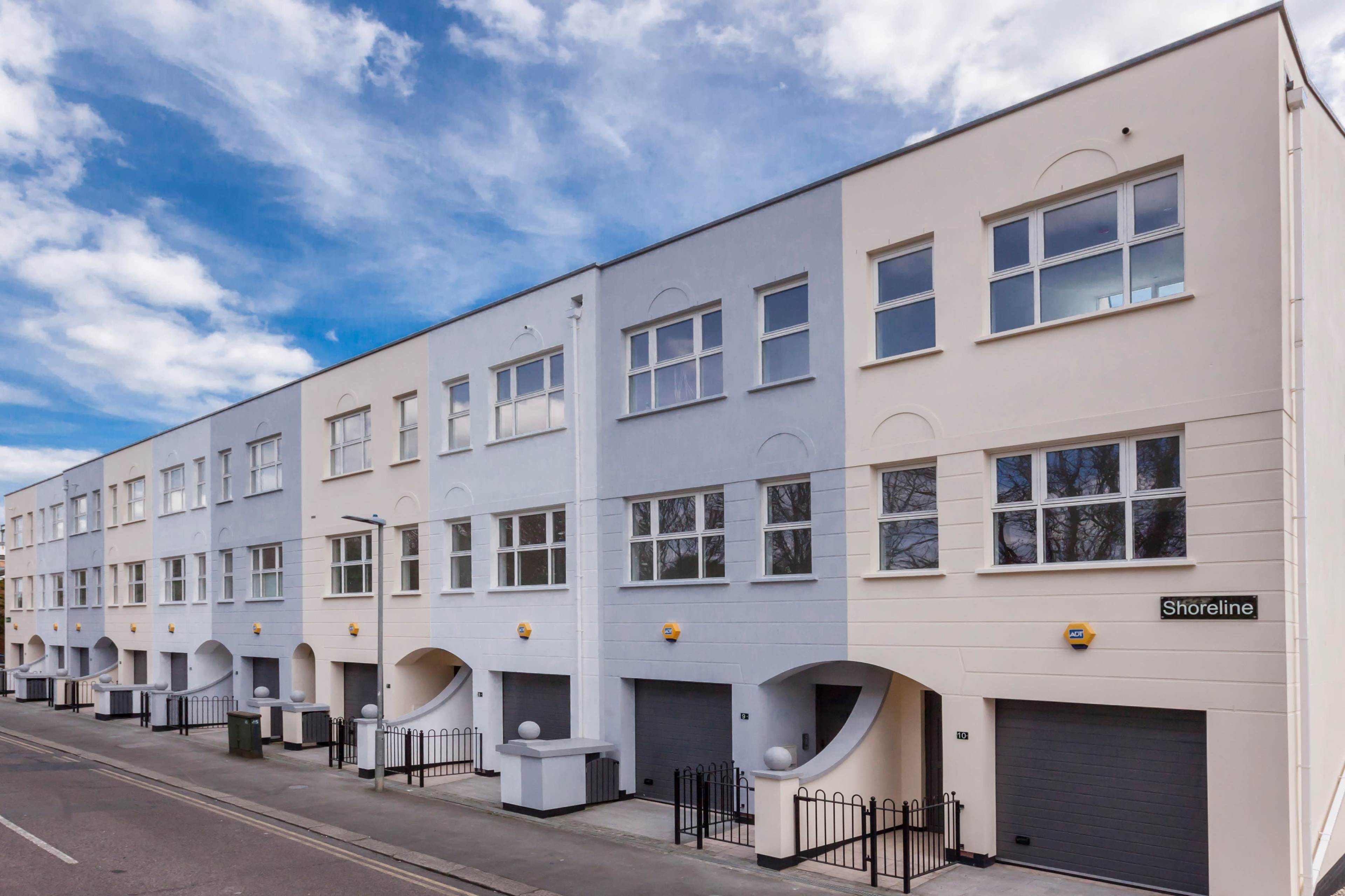A row of modern residential buildings with large windows and garage doors line a street under a blue sky.