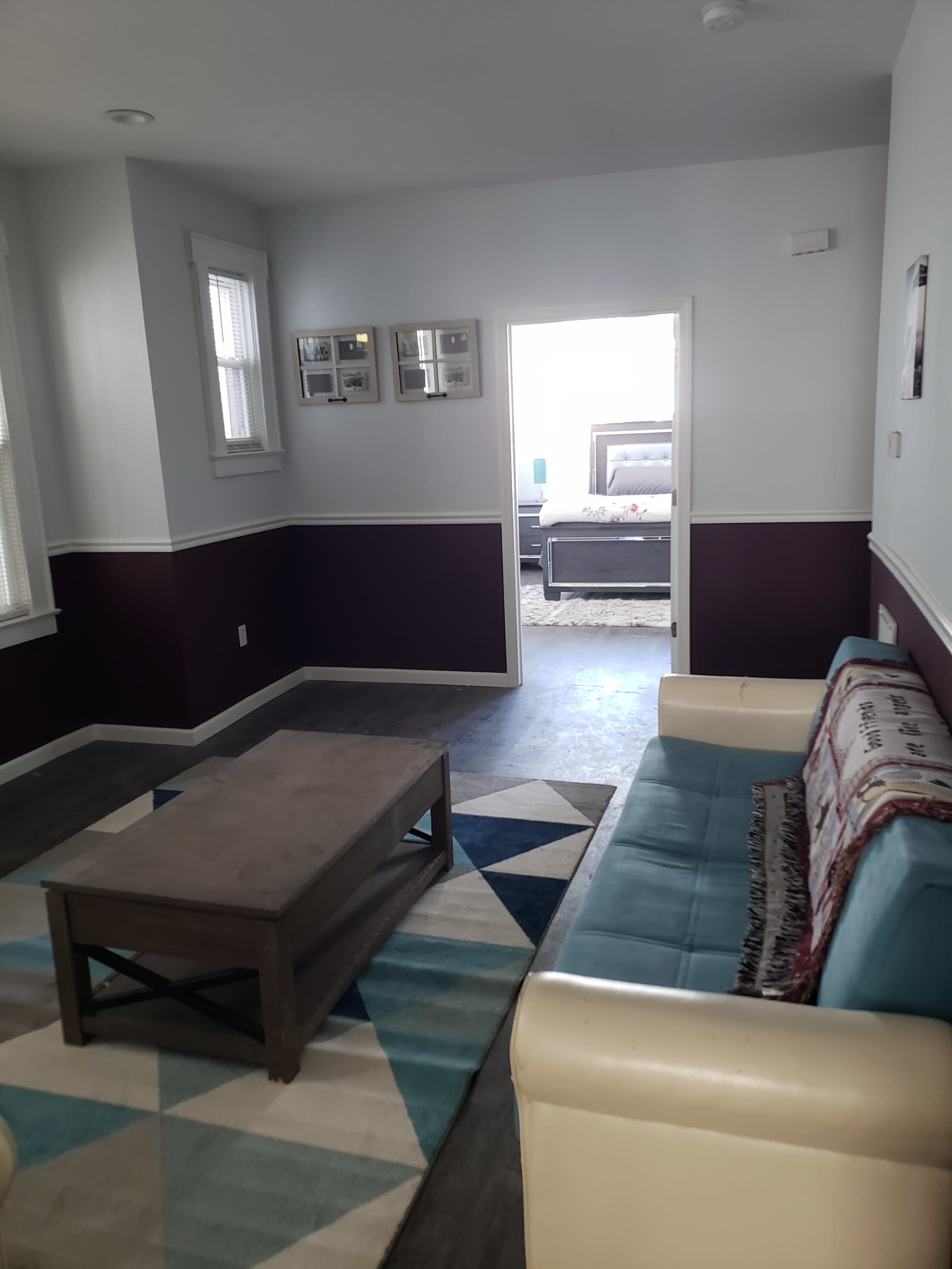 A living room featuring a teal sofa, a coffee table, and a geometric rug, with a doorway leading to a bedroom in the background.