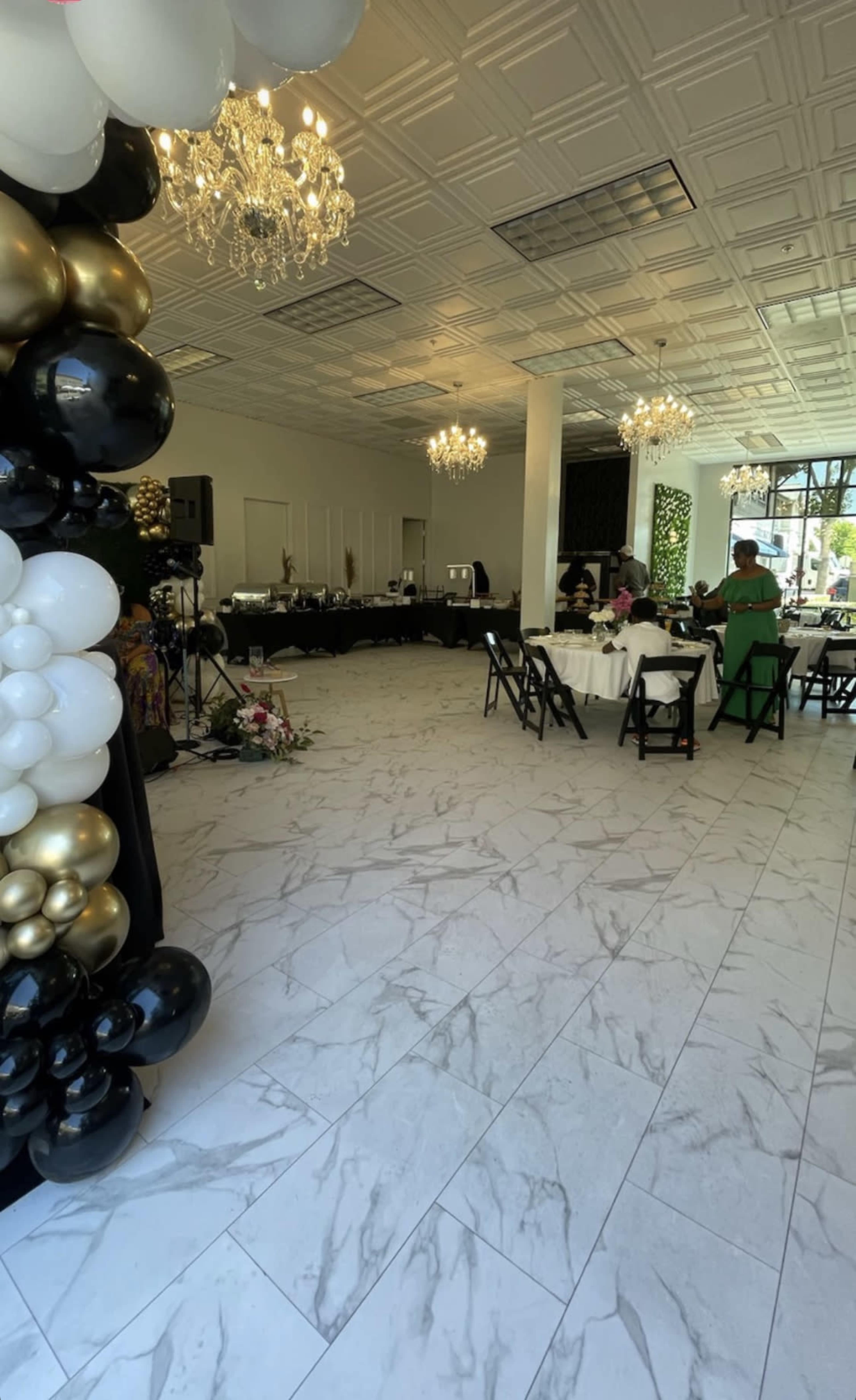 A spacious event venue with white marble floors, decorated with a black and gold balloon arch, and tables set for dining under chandeliers.