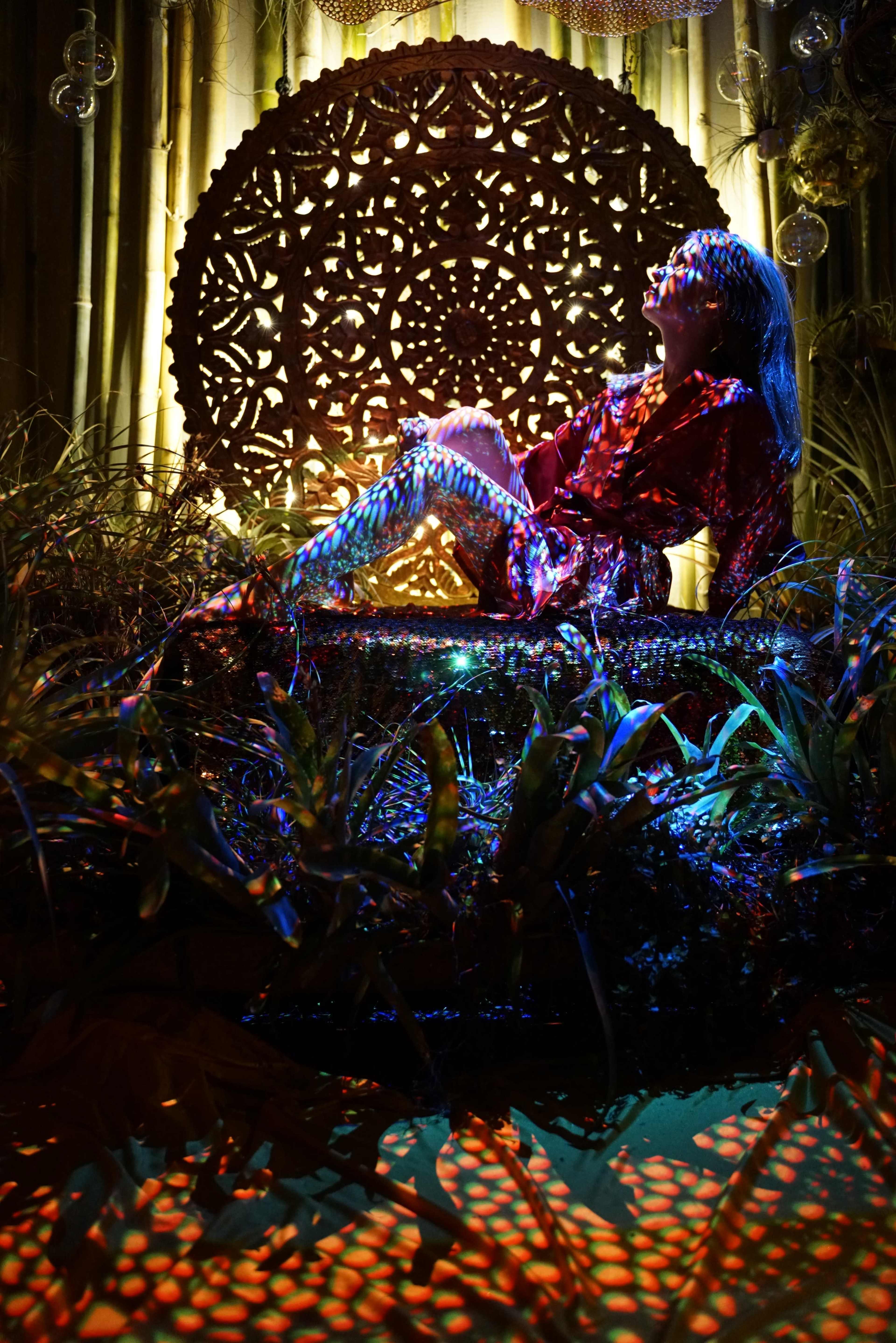 A figure in a red outfit sits against an ornate background, surrounded by vibrant lighting and lush greenery.