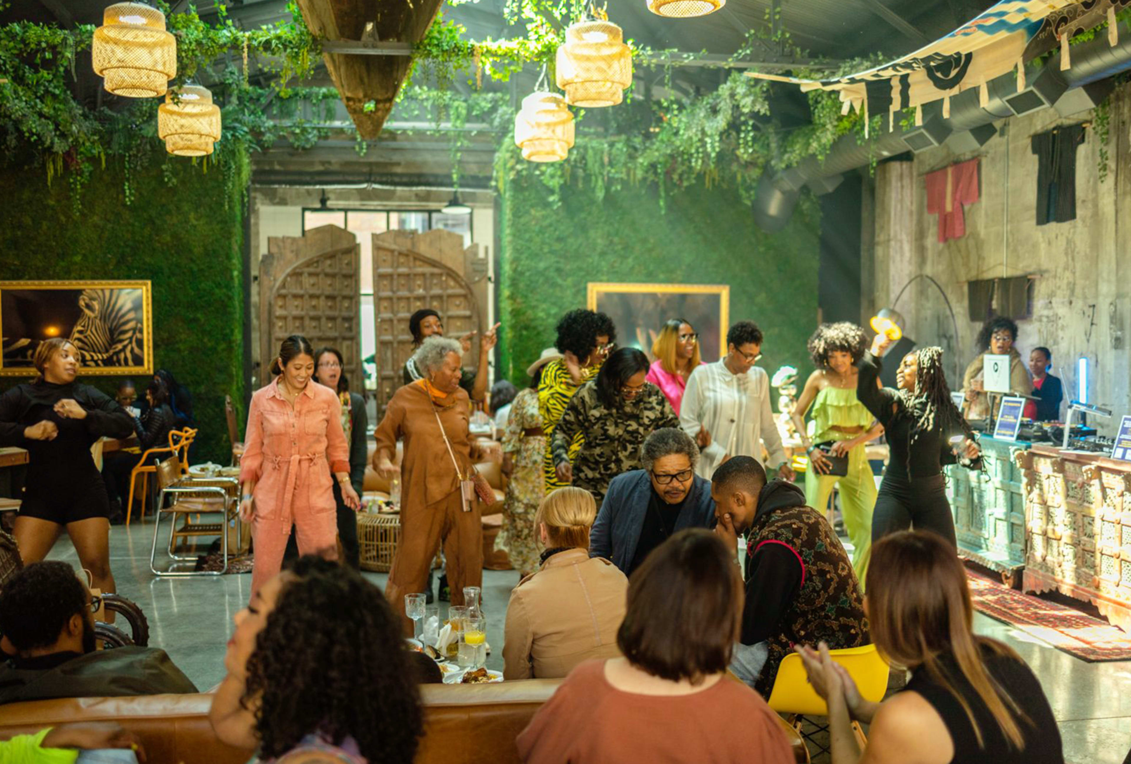 A lively gathering takes place in a green-themed venue with people dancing and socializing amid plants and artistic decor.
