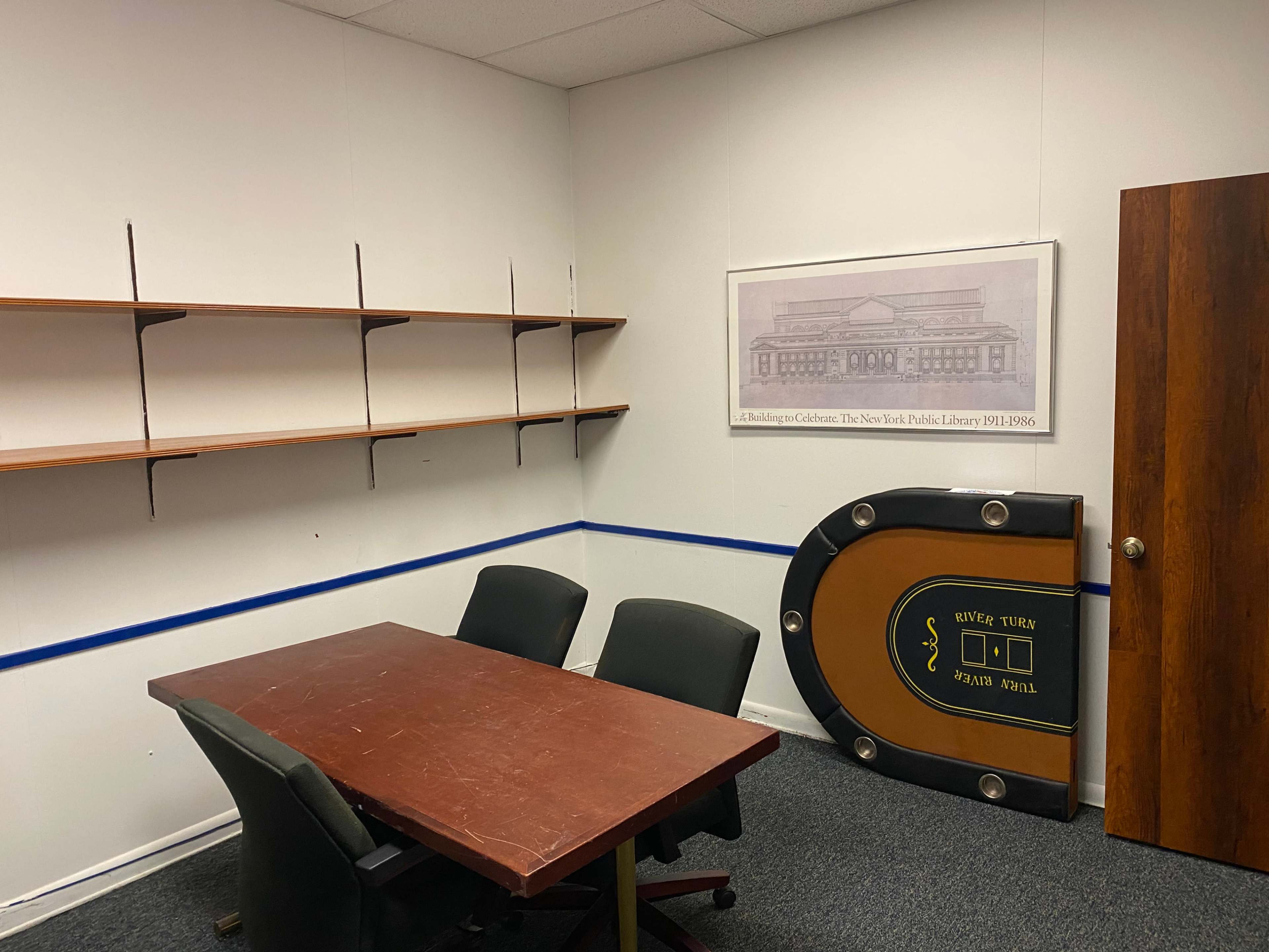 The image shows a small, empty office space with a wooden table and chairs, a set of shelves along the wall, and a framed artwork depicting a library.