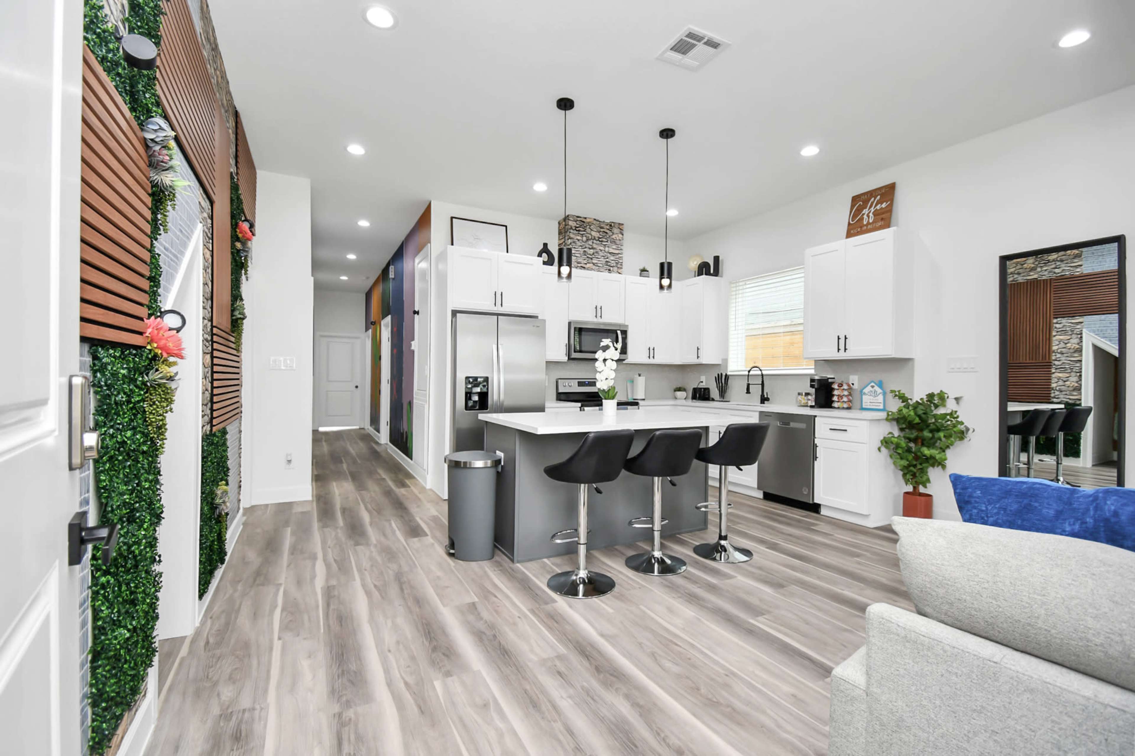 A modern kitchen features white cabinets, a central island with black stools, and wood-style flooring, illuminated by pendant lights.