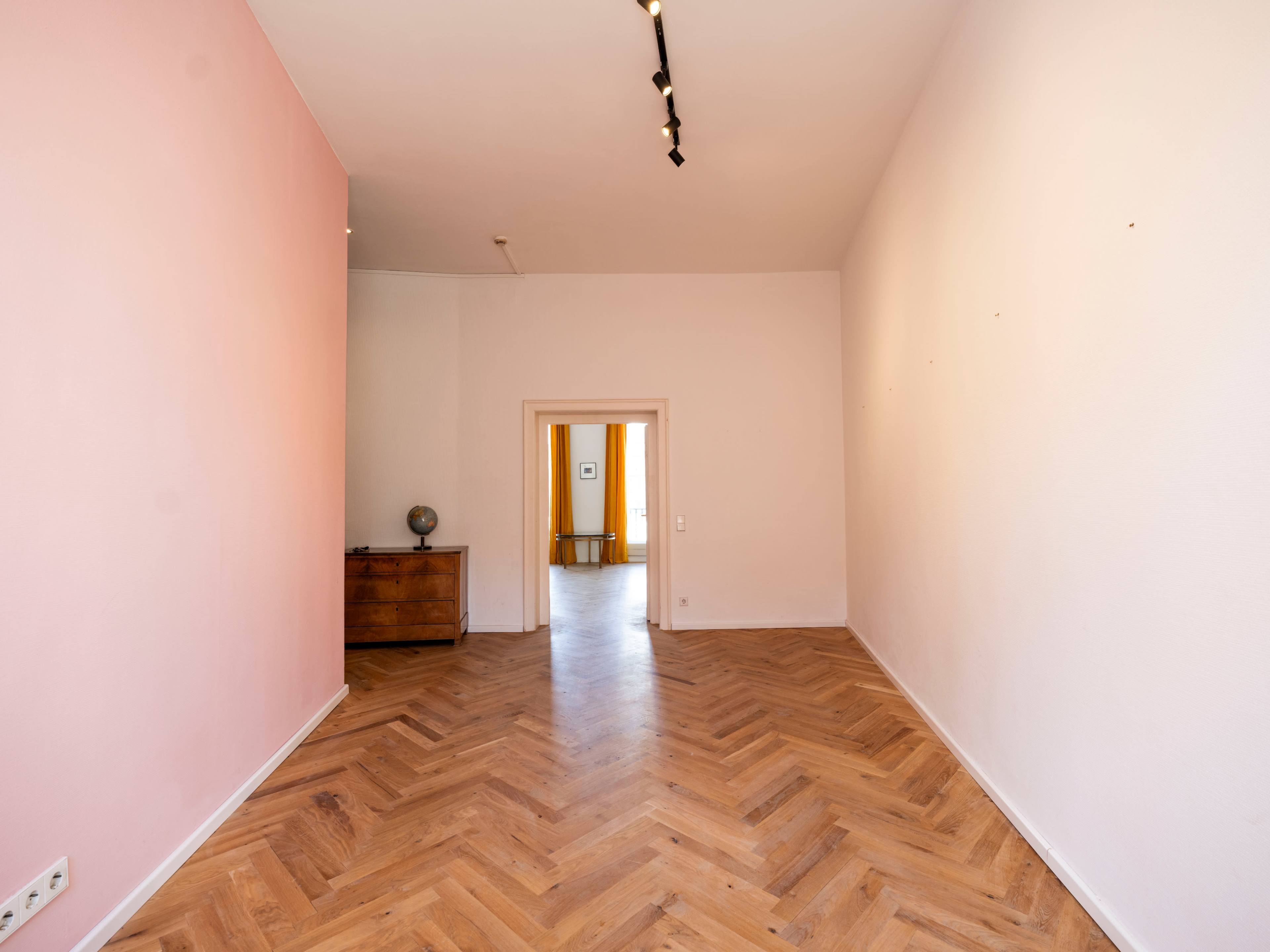 The image shows an empty room with light pink walls and wooden herringbone flooring leading to an open doorway.