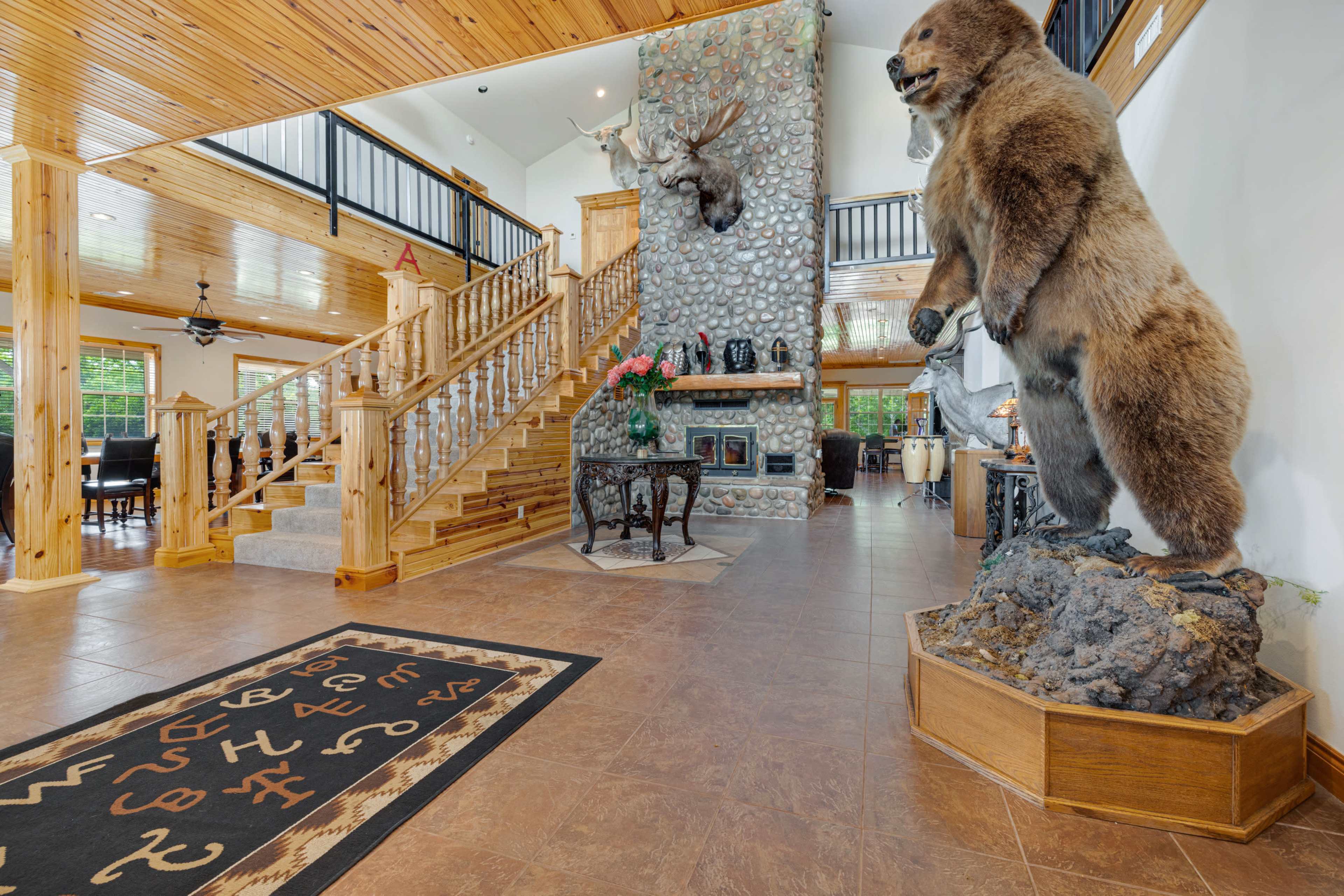 The entrance features a grand staircase, a stuffed bear mount, and a stone fireplace surrounded by wooden accents.