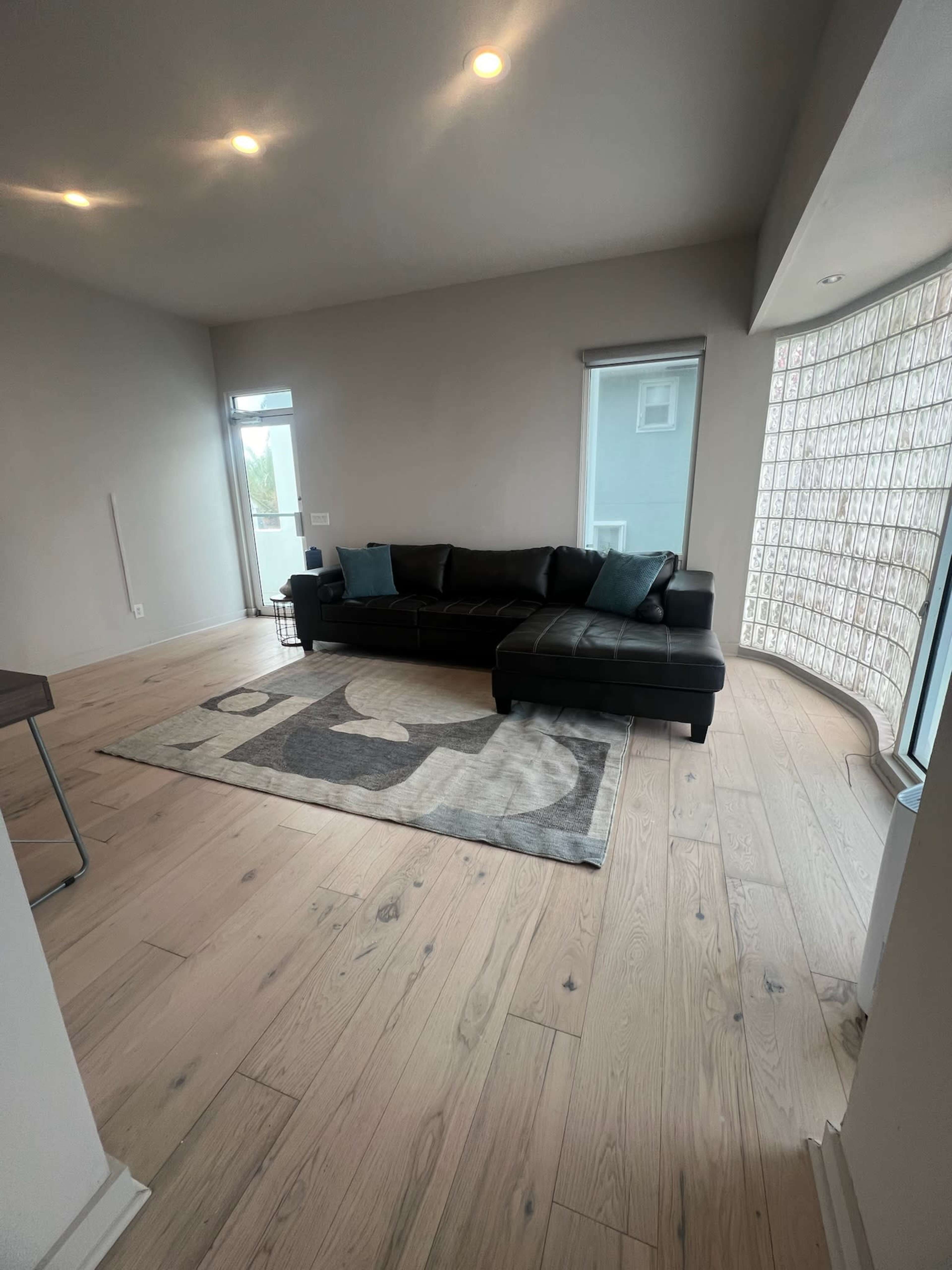 The image shows a light-filled living room featuring a black sectional sofa, a patterned area rug, and large windows with glass block accents.