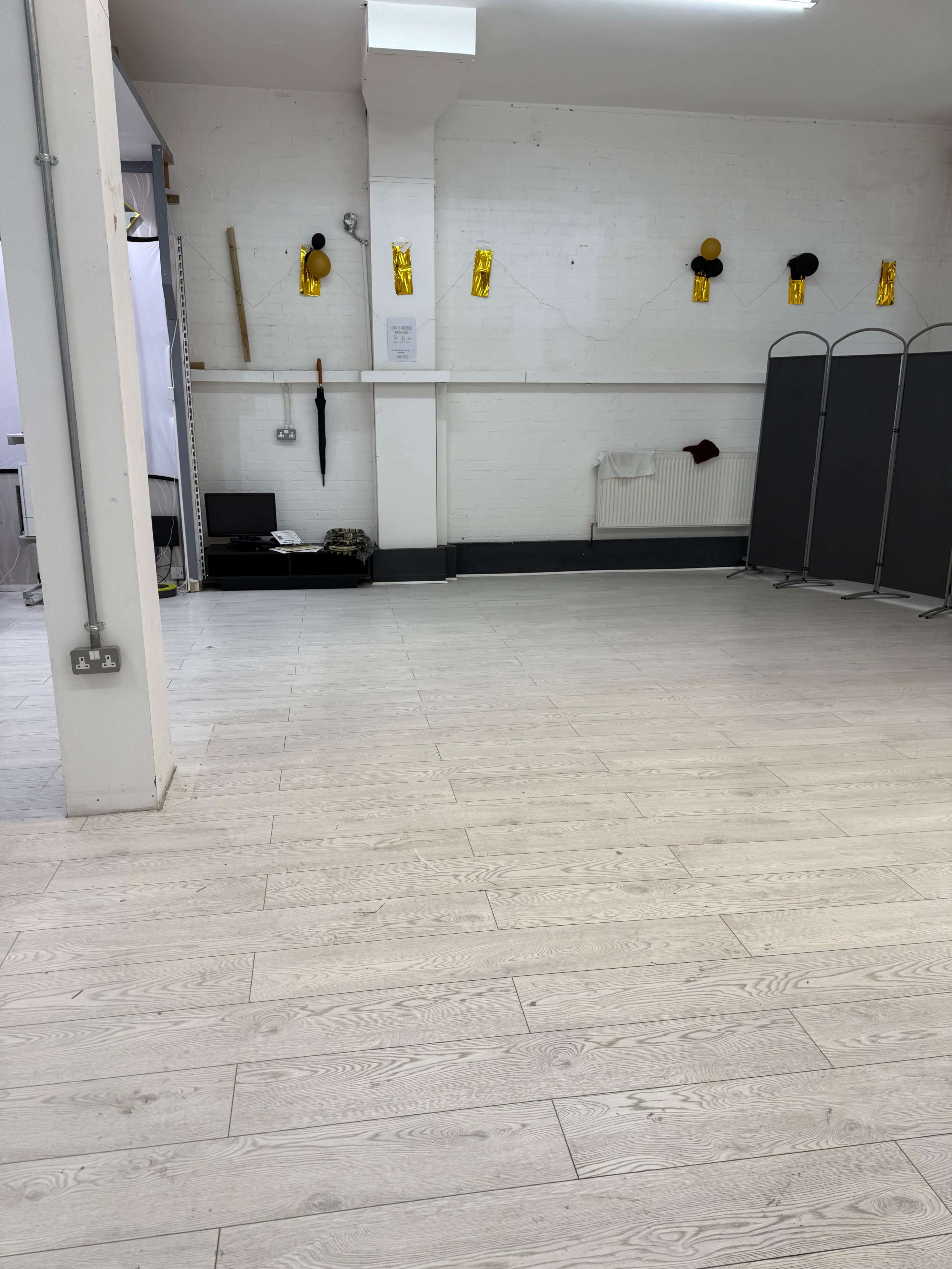 The image shows a spacious, empty room with light-colored wooden flooring, white brick walls, and a few simple partitions along the back.