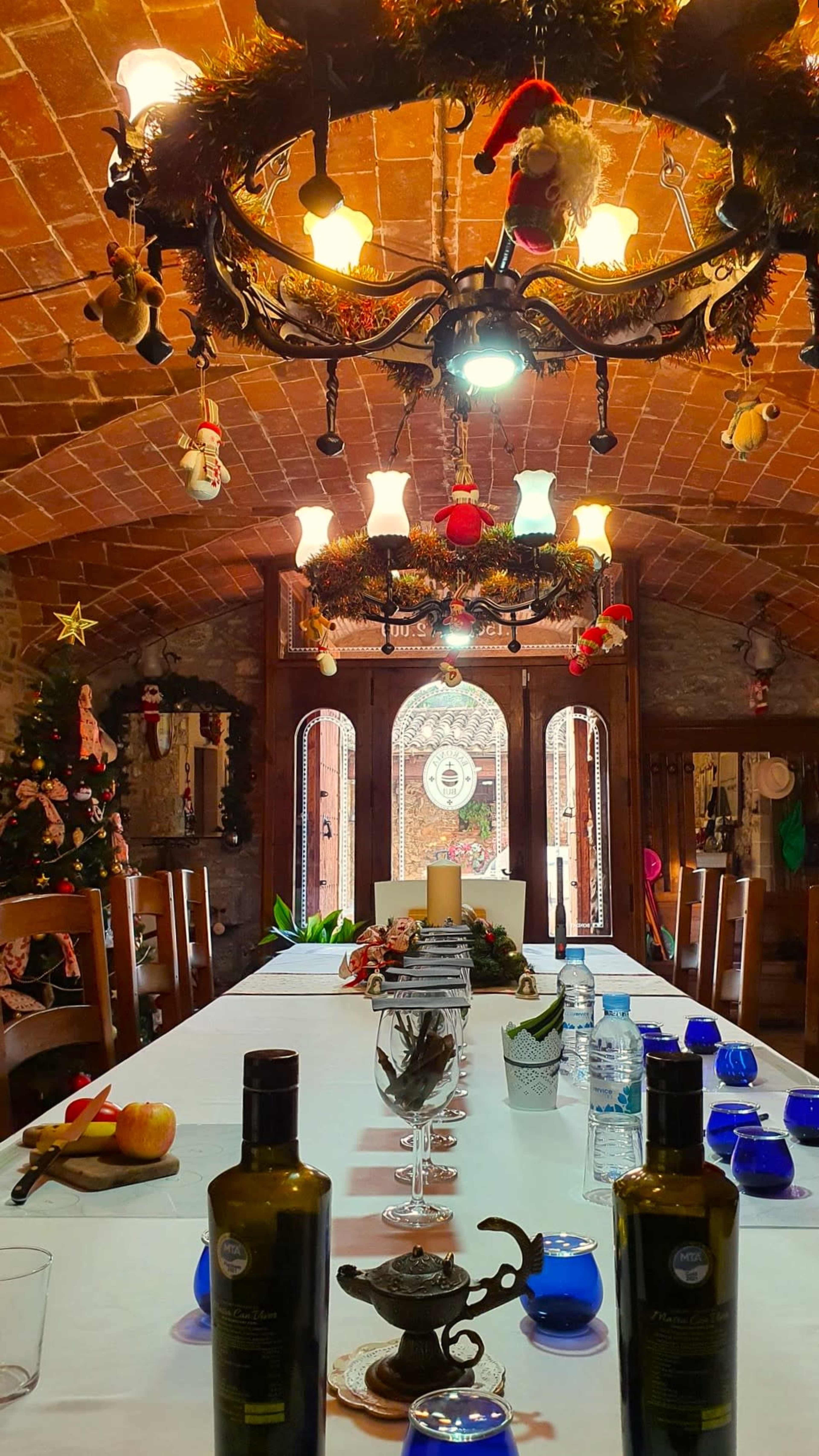 A long dining table is set in a rustic restaurant decorated with a Christmas tree and festive ornaments hanging from the chandelier.