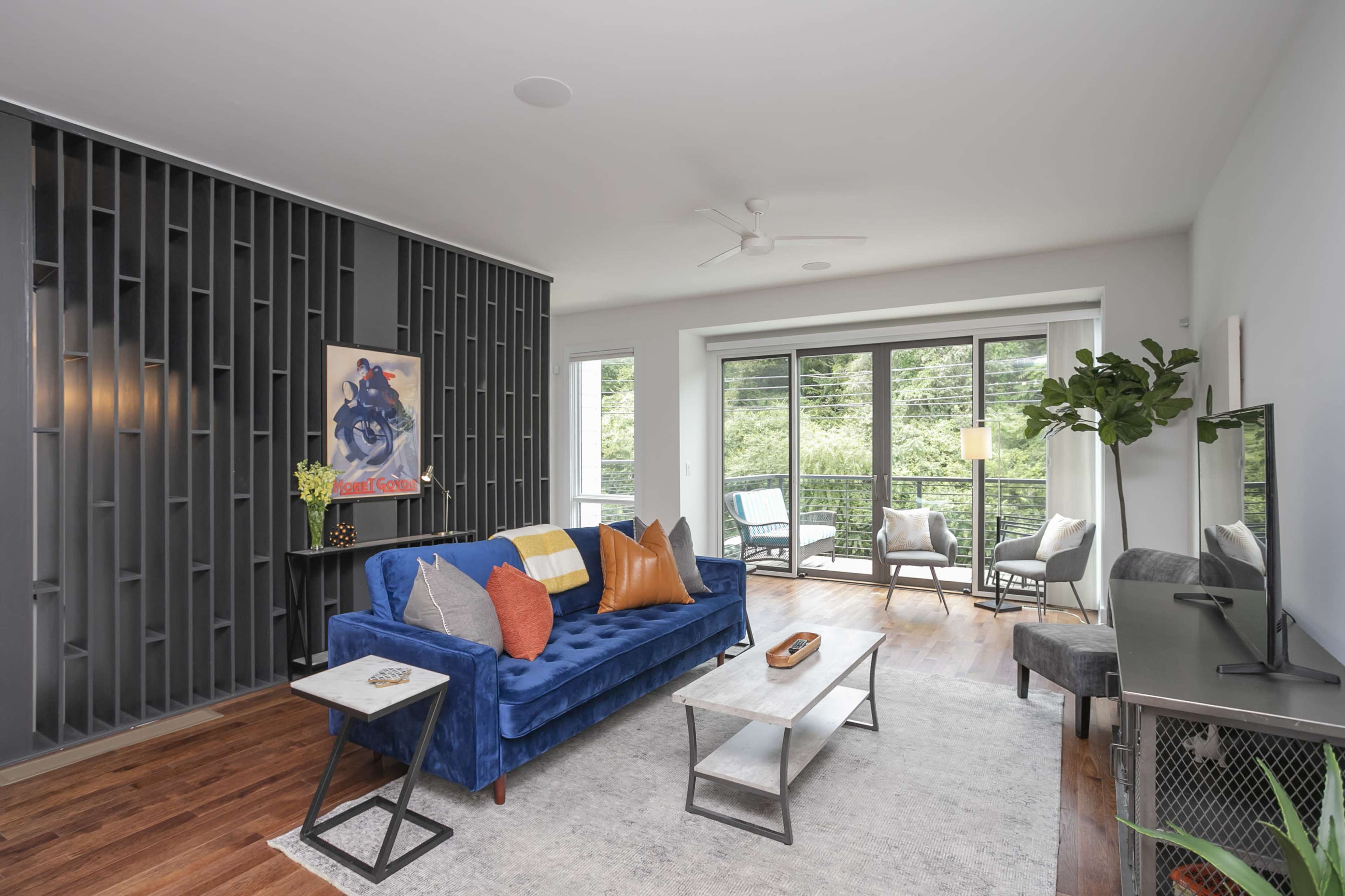 A modern living room features a blue sofa, a gray coffee table, and large windows overlooking a green landscape.