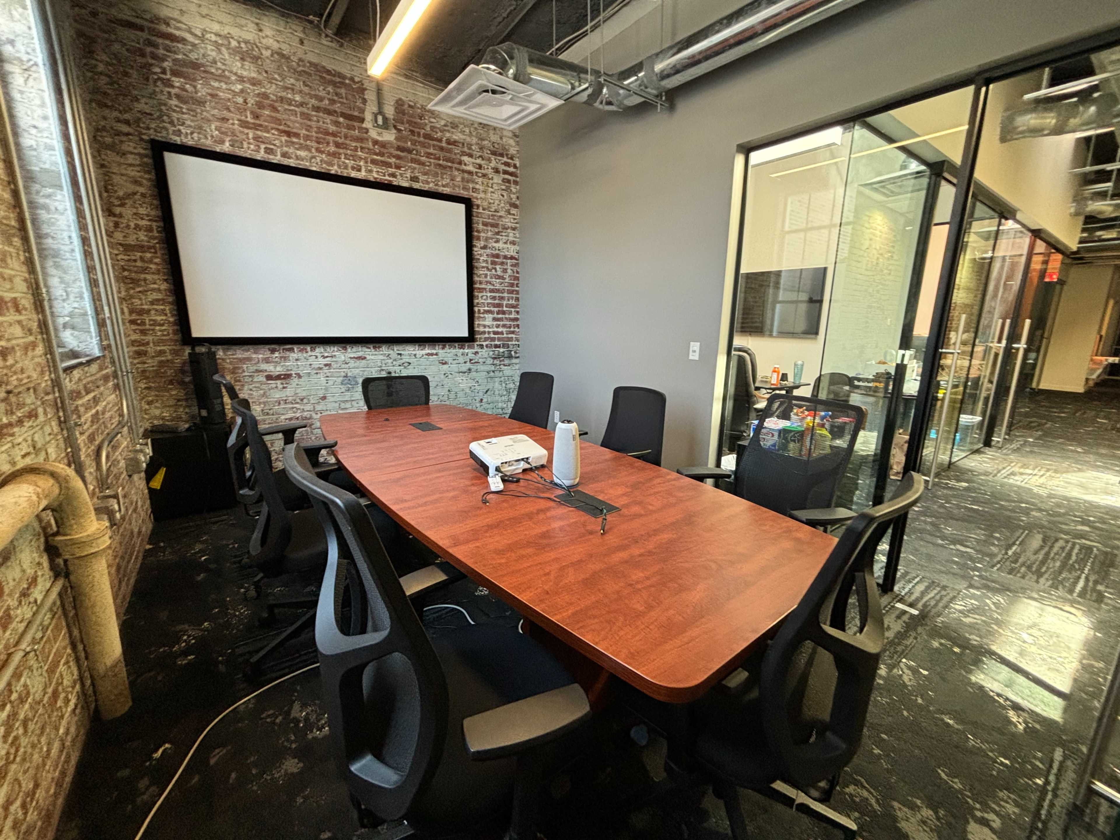 A conference room with a large wooden table surrounded by black chairs, a projector on the table, and a screen mounted on the wall.