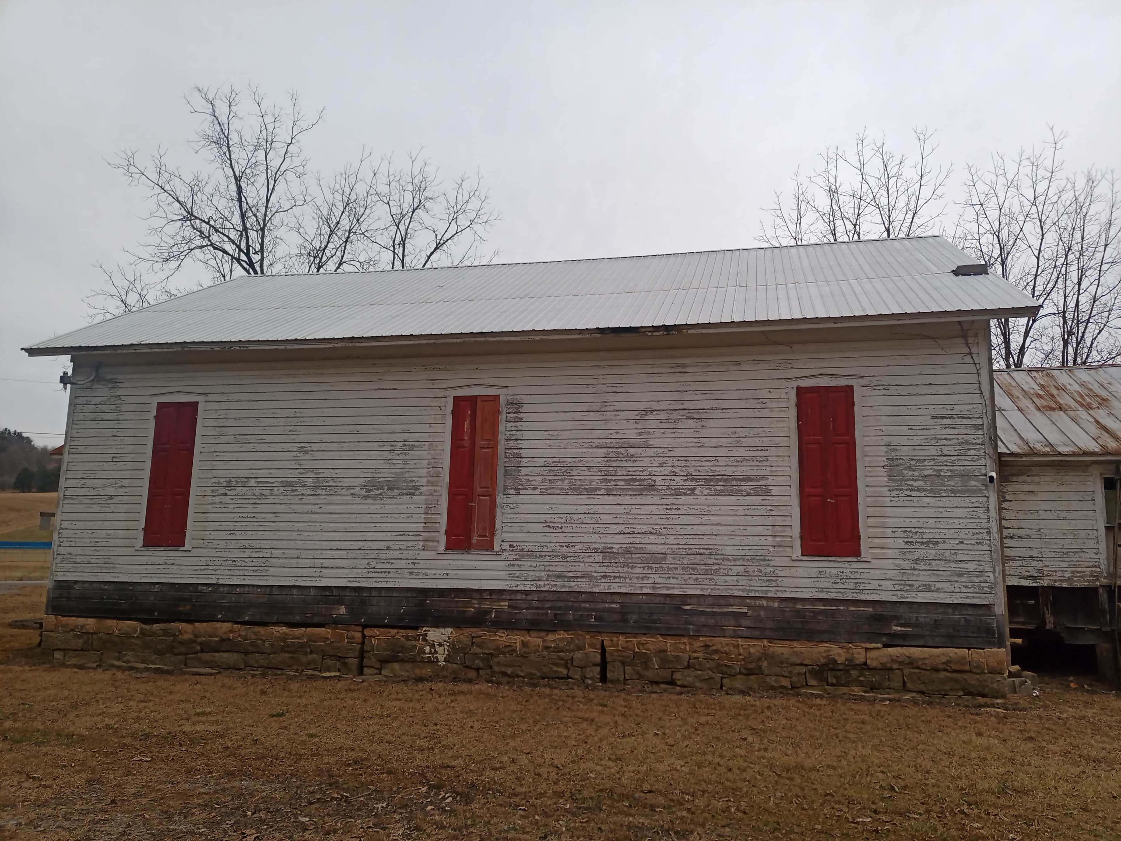 A weathered wooden building with a metal roof features red shutters on its sides, situated on grassy ground.