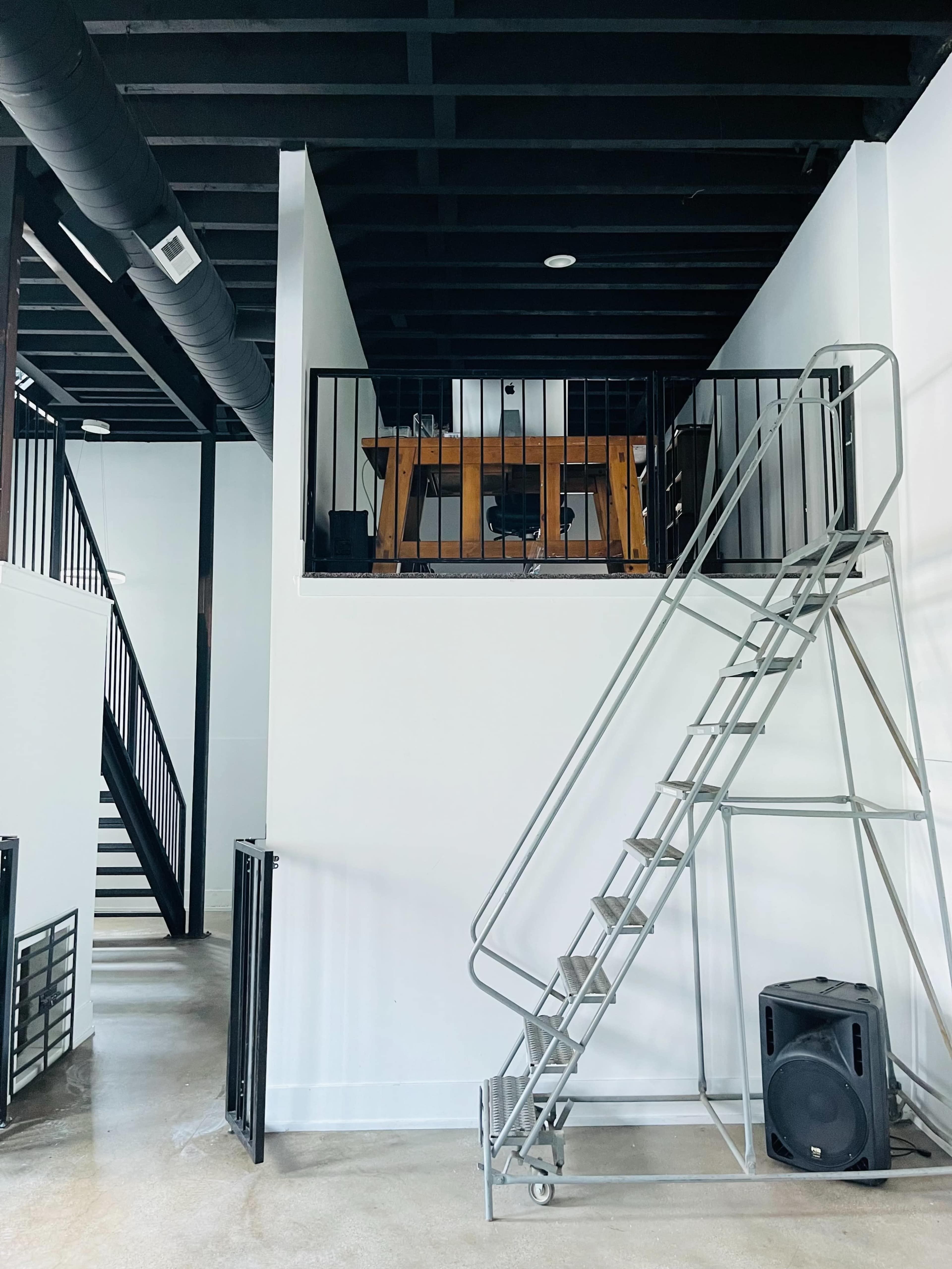 A modern interior with a metal staircase leading to a raised loft area that features a wooden table and chair, surrounded by black metal railings.