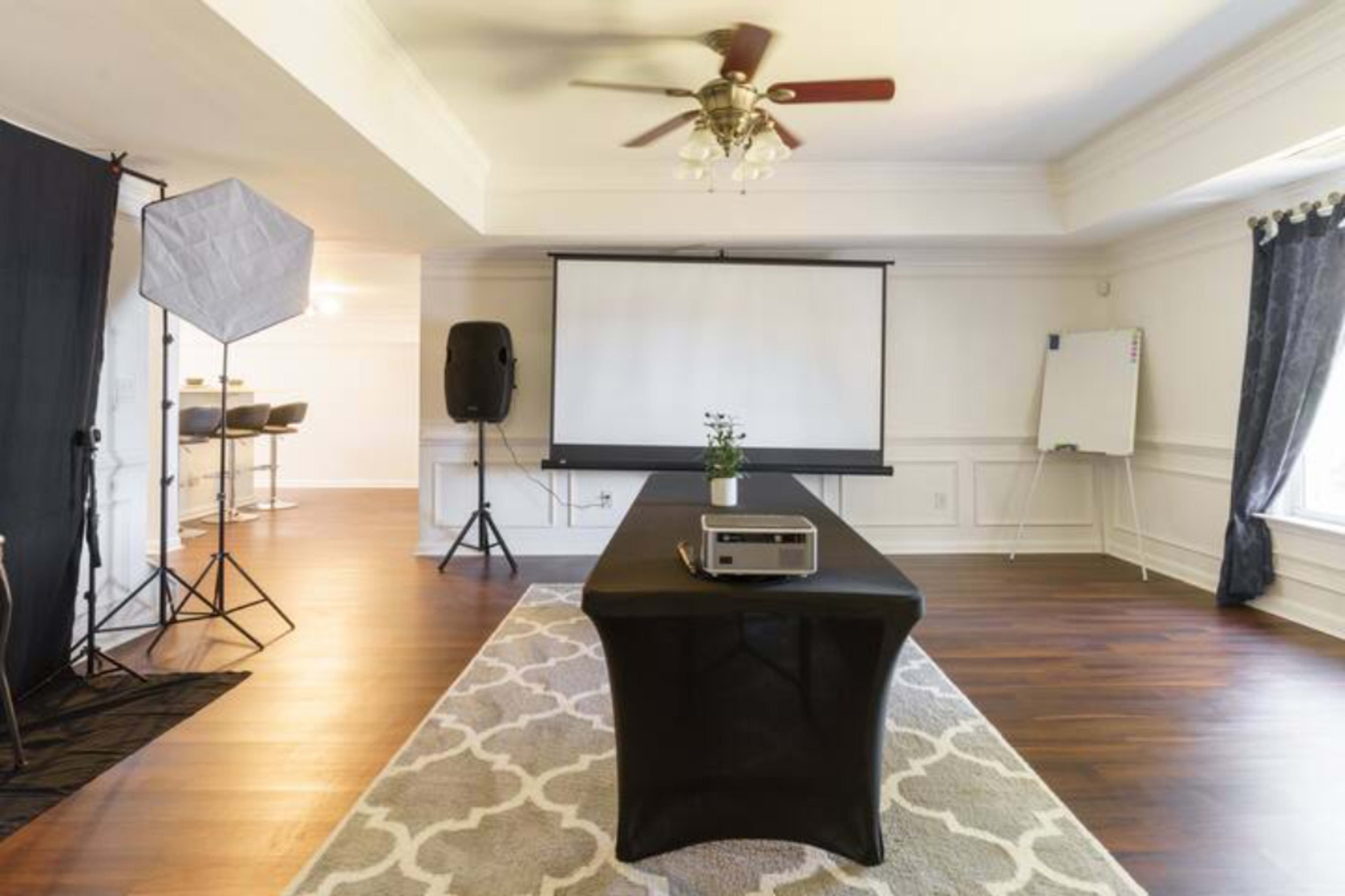 A spacious meeting room features a central black table with a projector, a large screen at the front, and two additional setups in the background for audio and visual equipment.